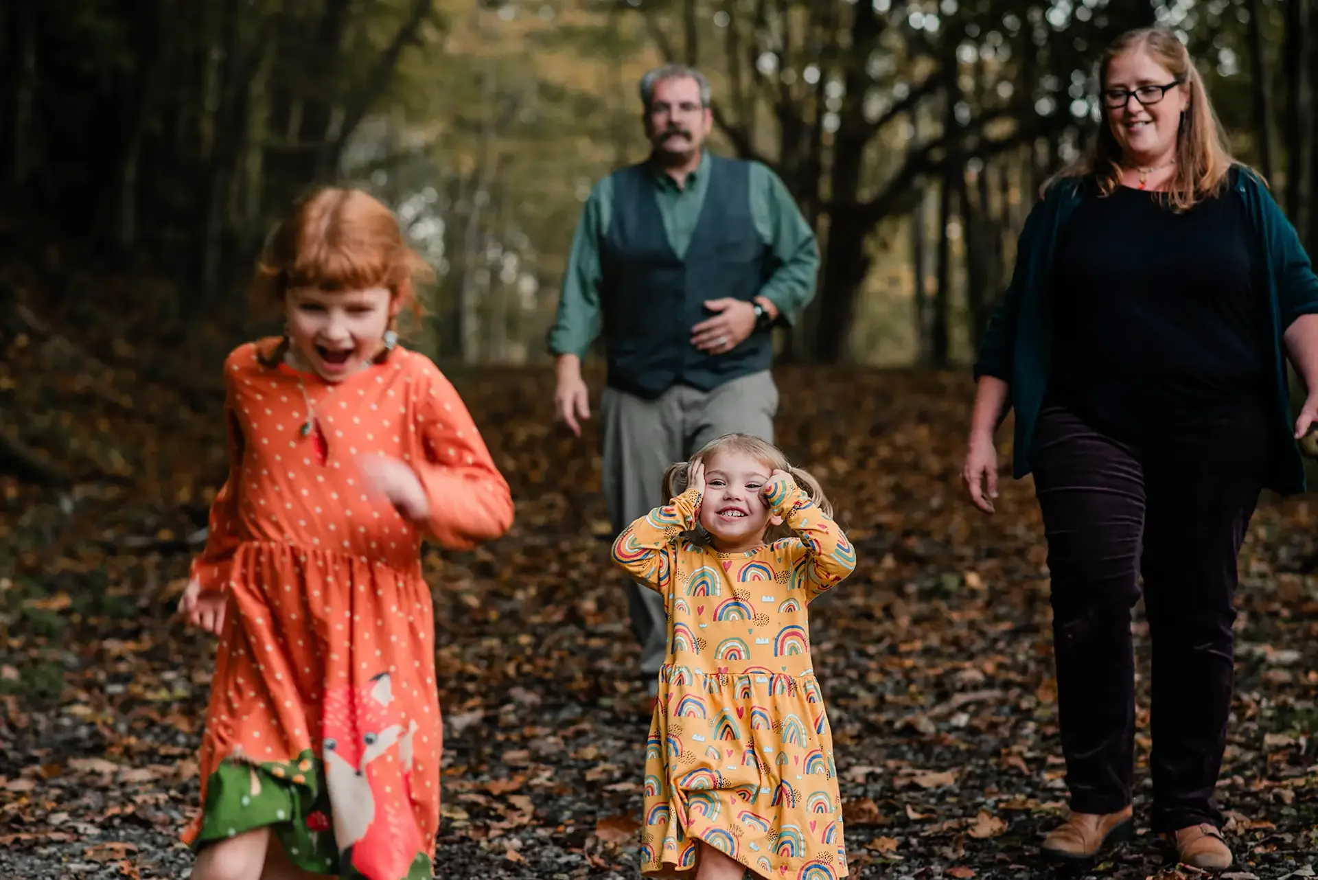 A family of four - mom, dad, and two sisters - walk through a forest path in the fall, they are dressed warmly. The youngest holds her hands to her head like "can you believe these people?"