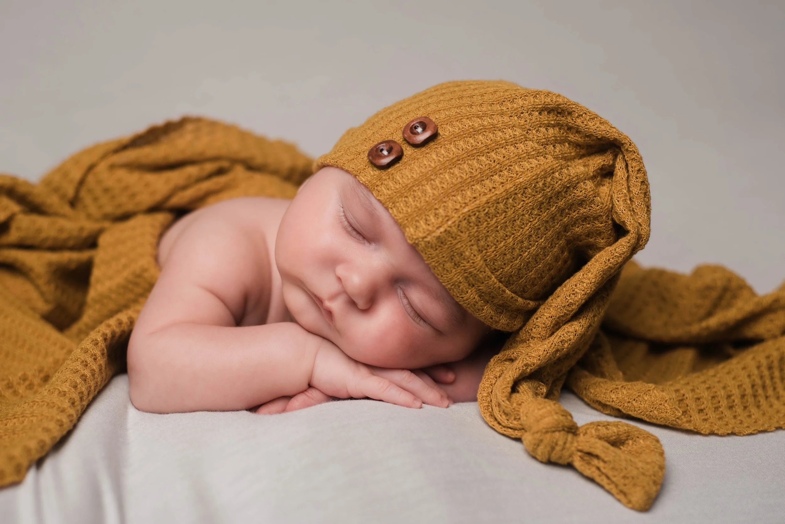 A sleeping baby with a mustard yellow knit hat and blanket, resting on their side with hands under their chin. Studio newborn portrait, Brownsville PA.