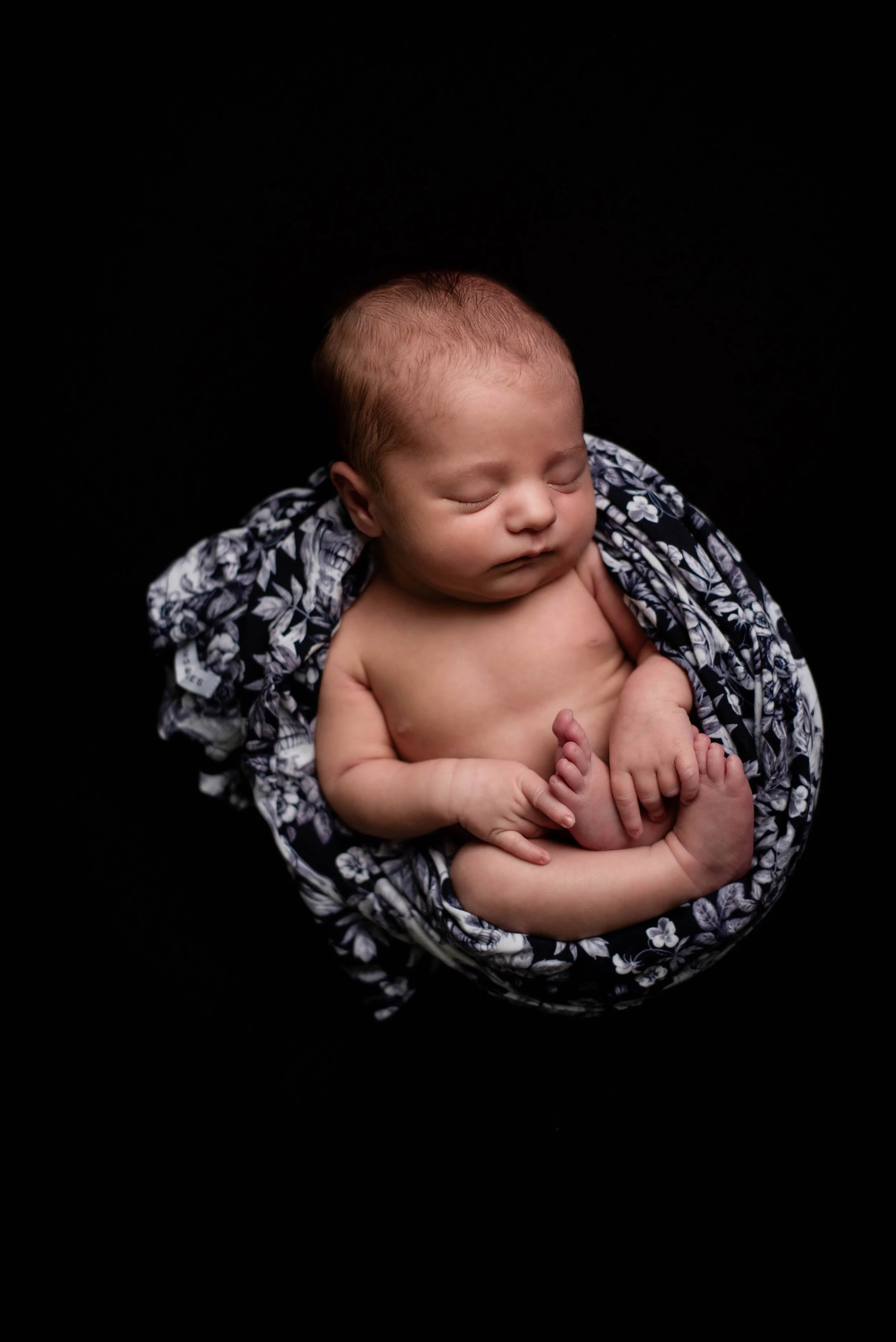 A sleeping newborn baby wrapped in a floral blanket with skulls against a black background. Studio newborn portrait, Brownsville PA.