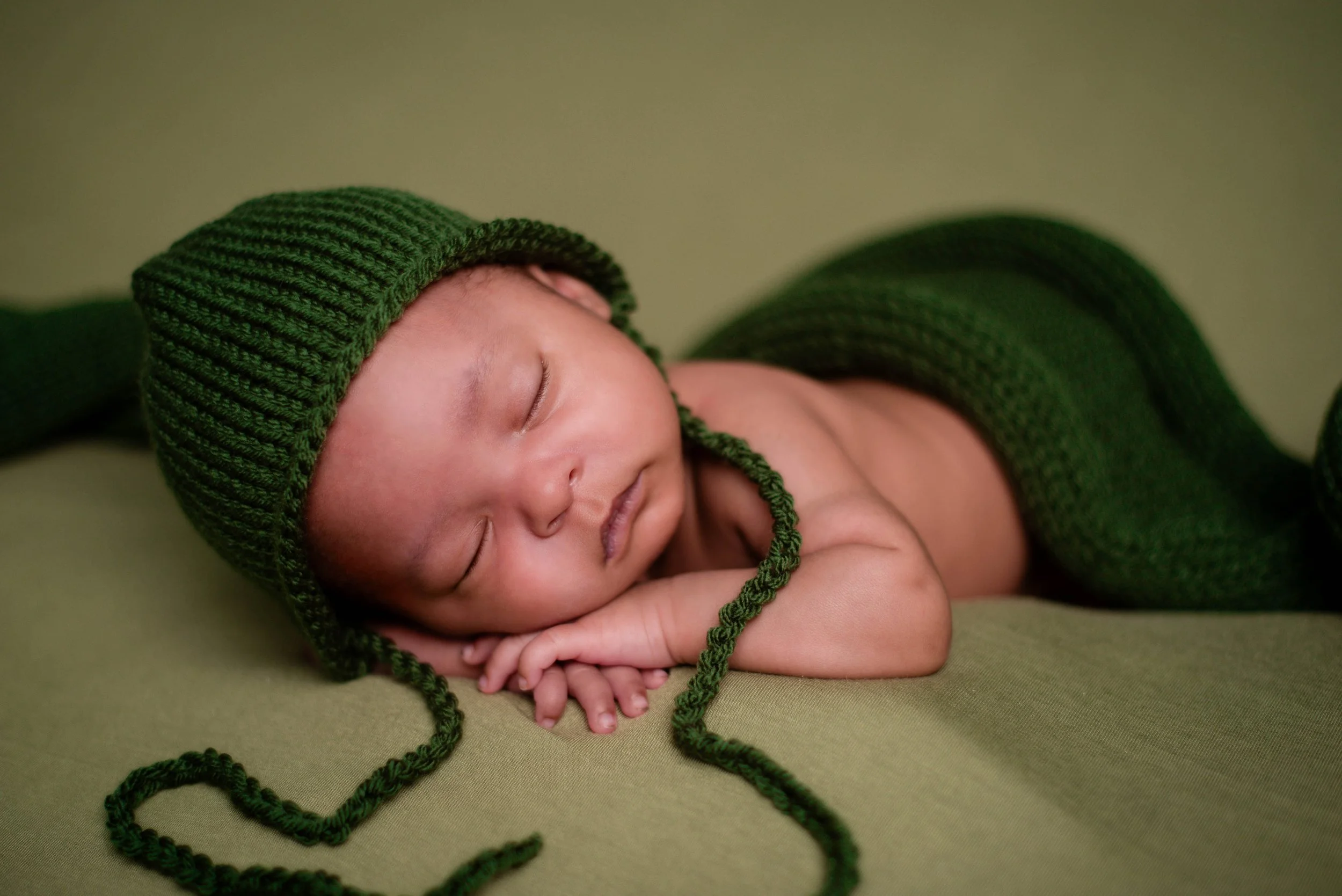 A sleeping baby wearing a green knitted hat and wrapped in a matching green blanket, lying on a green surface. Studio newborn portrait, Brownsville PA.