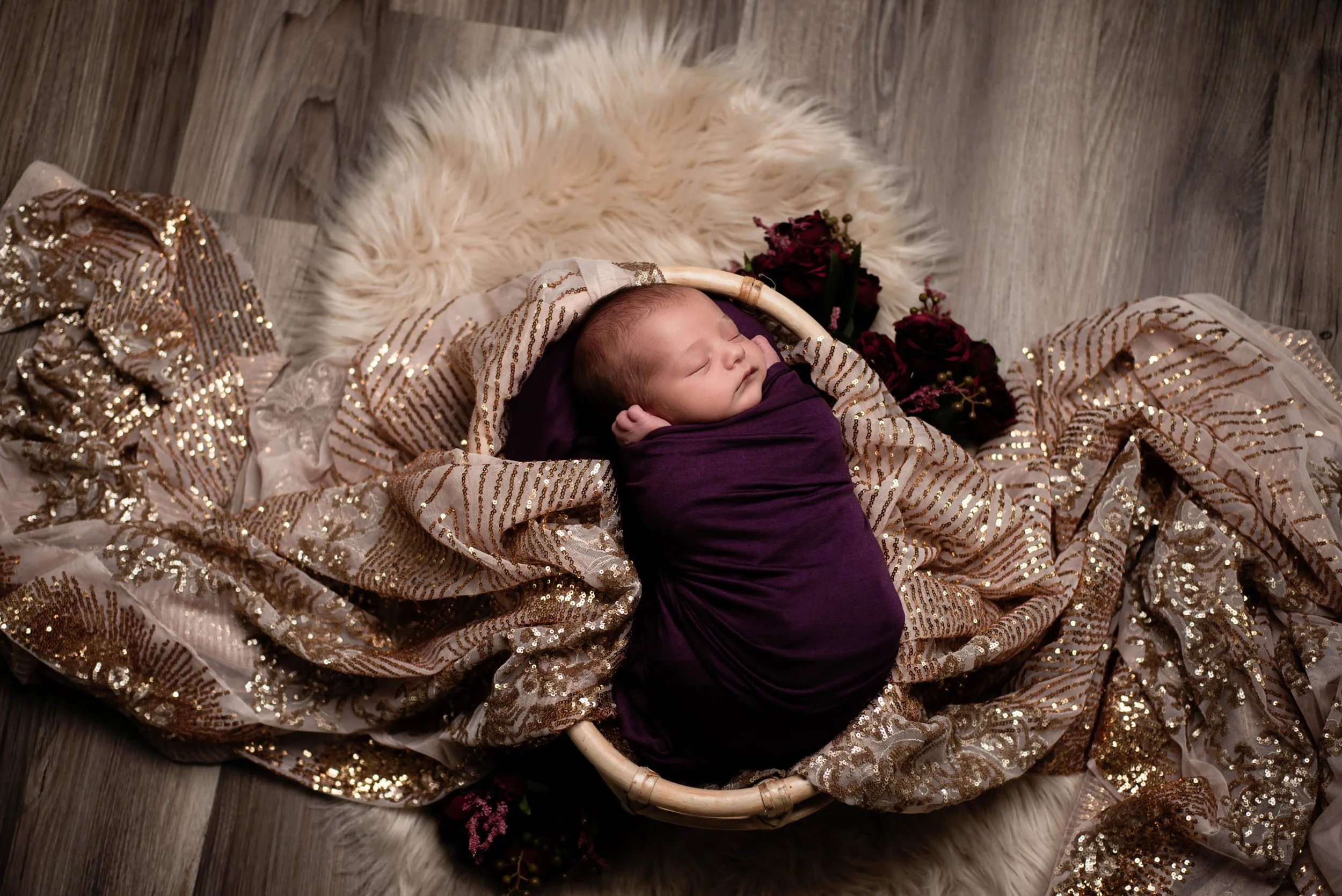A newborn baby is sleeping in a basket, wrapped in a purple blanket, surrounded by shimmering gold and silver fabric, with dark red roses beside the basket, on a fluffy white rug over wooden floor. Studio newborn portrait, Brownsville PA.