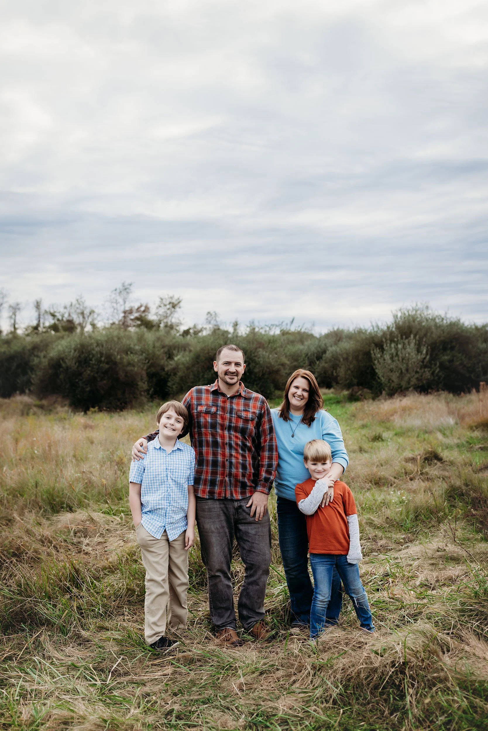 A family of four standing outdoors in a grassy field with bushes and trees in the background, under an overcast sky. Photographer Caroline Moore and her family.