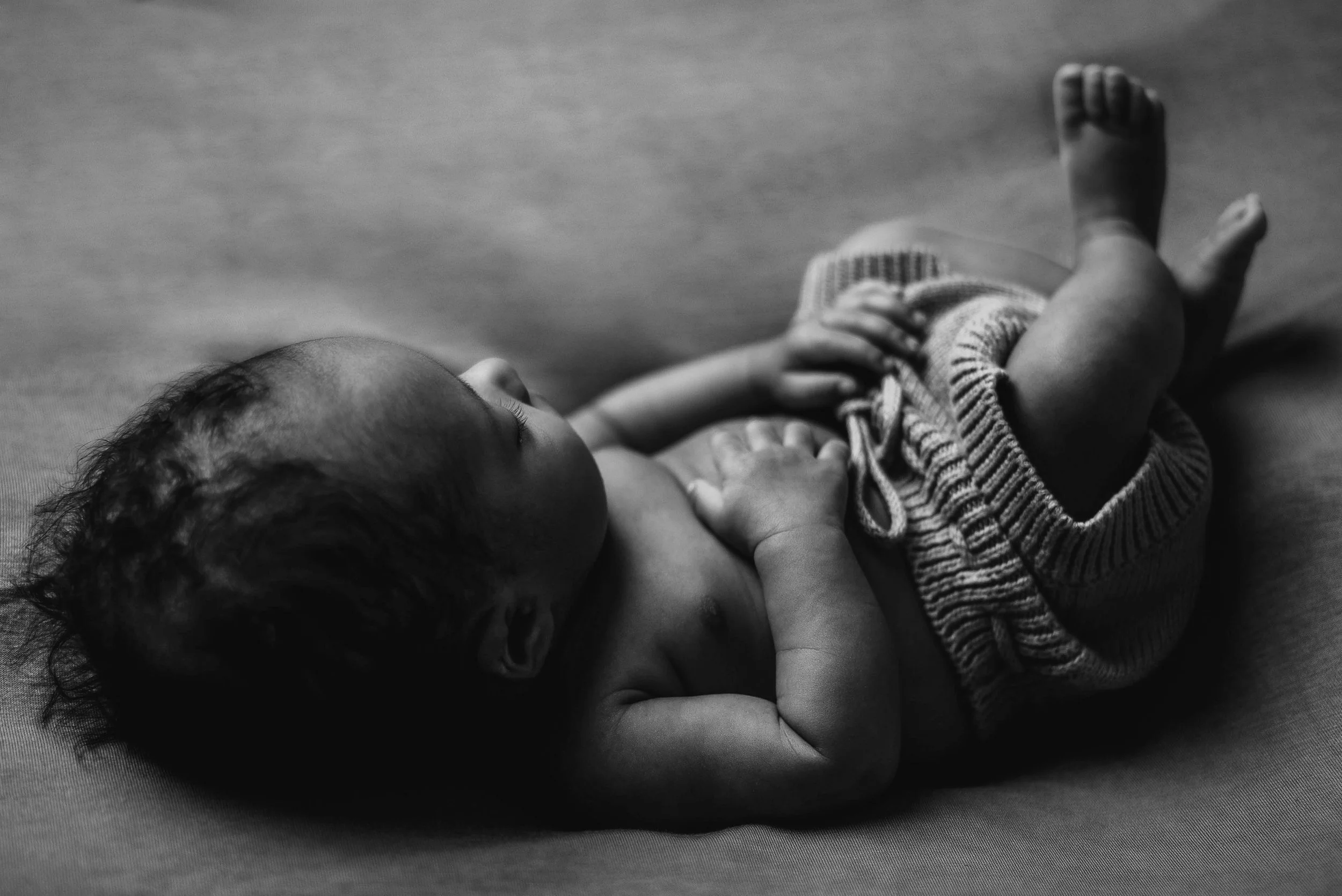 A black and white photo of a baby lying on its back on a soft surface, wearing shorts, with one hand resting on its chest and the other near its face. Studio newborn portrait, Uniontown PA.