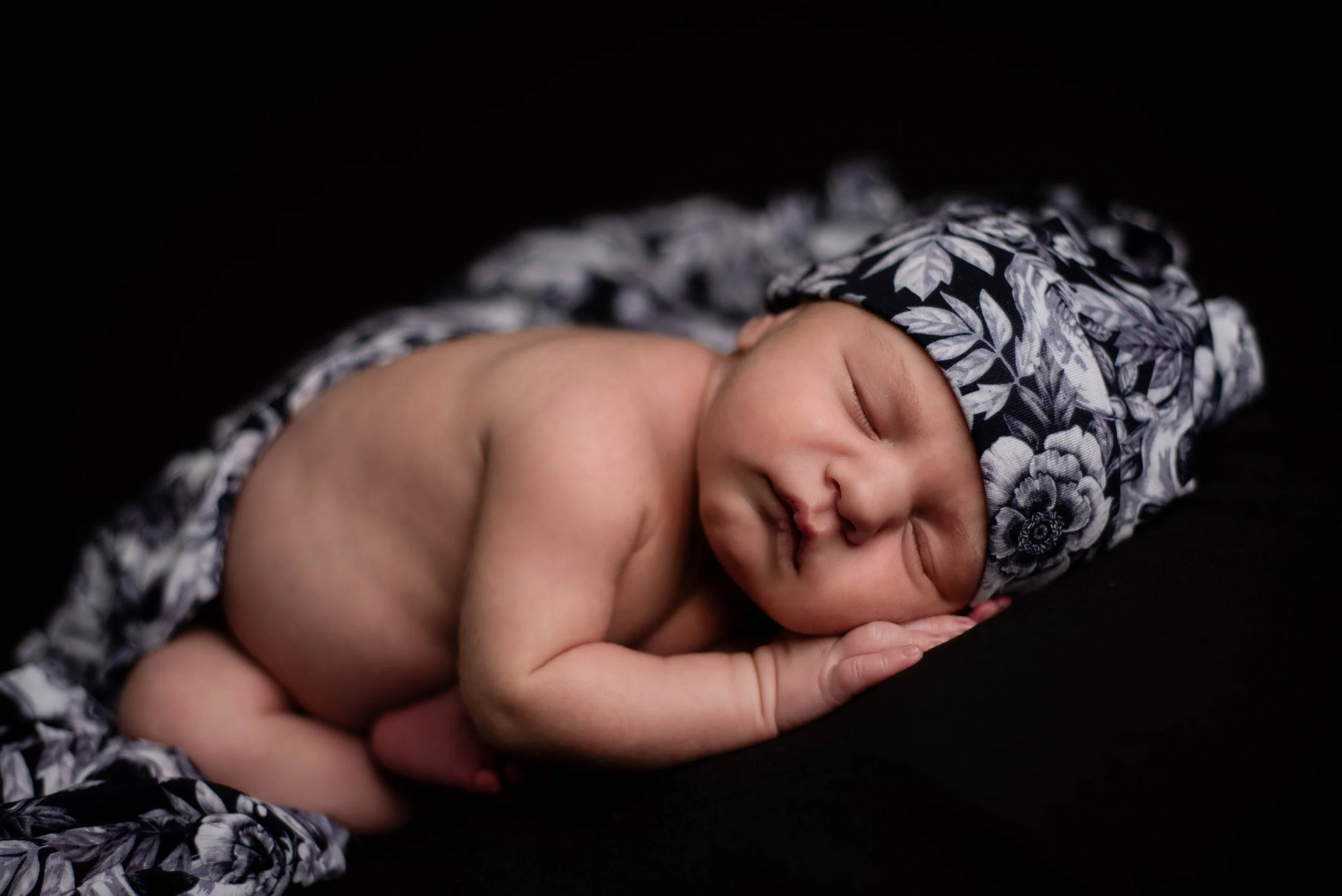 A sleeping newborn baby lying on a dark surface, wearing a black and white floral hat and blanket with skulls. Studio newborn portrait, Brownsville PA.