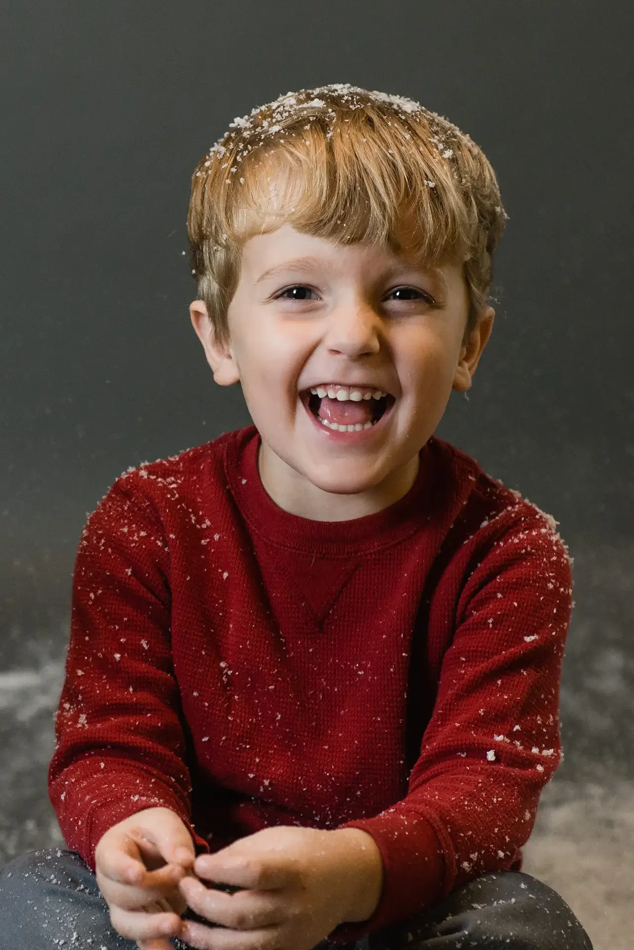 a young boy covered in fake snow for christmas snowglobe mini sessions, he is blonde and wearing a red sweater