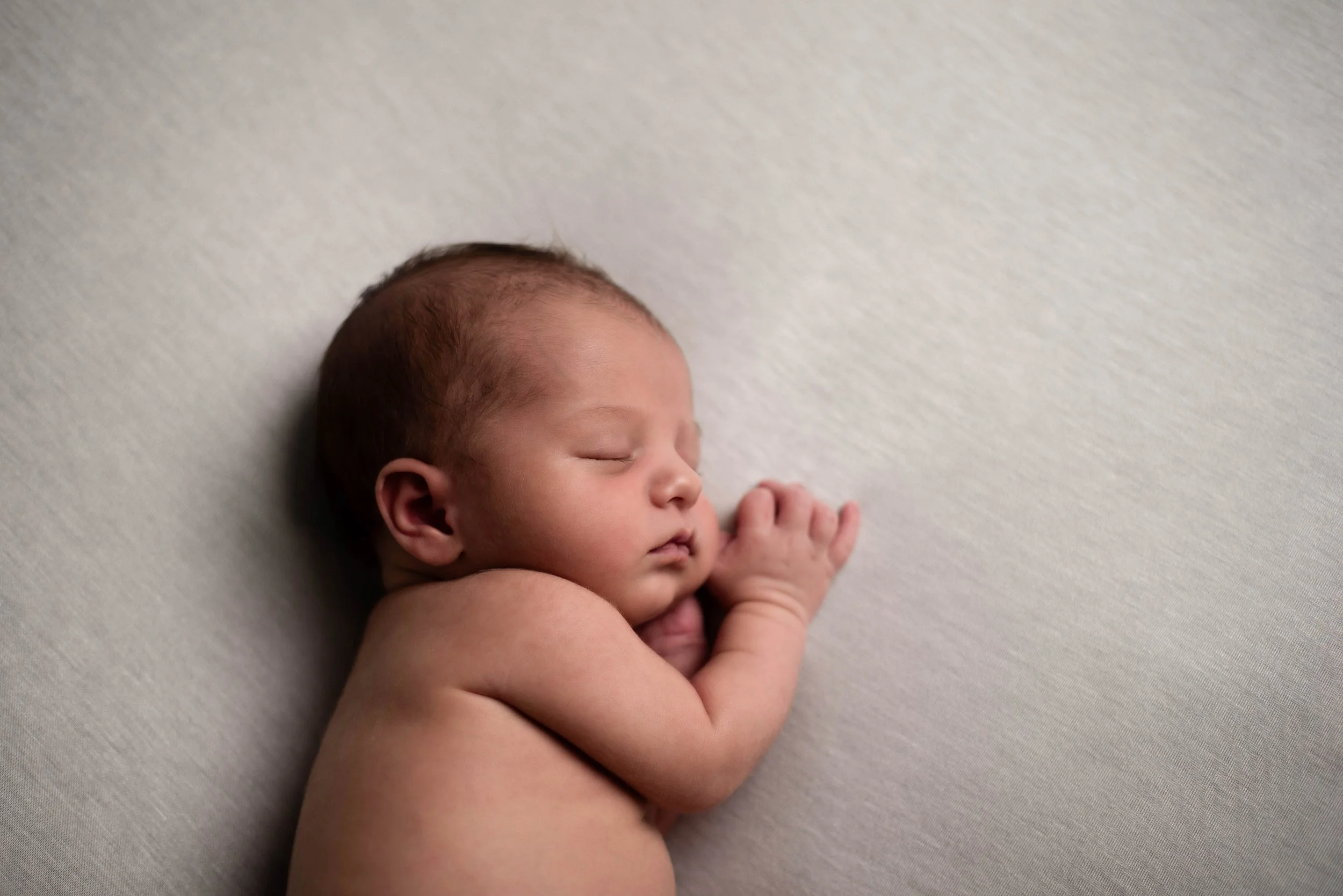 A sleeping newborn baby lying on a soft, light-colored surface, with eyes closed and tiny hands curled near the face. Studio newborn portrait, Brownsville PA.