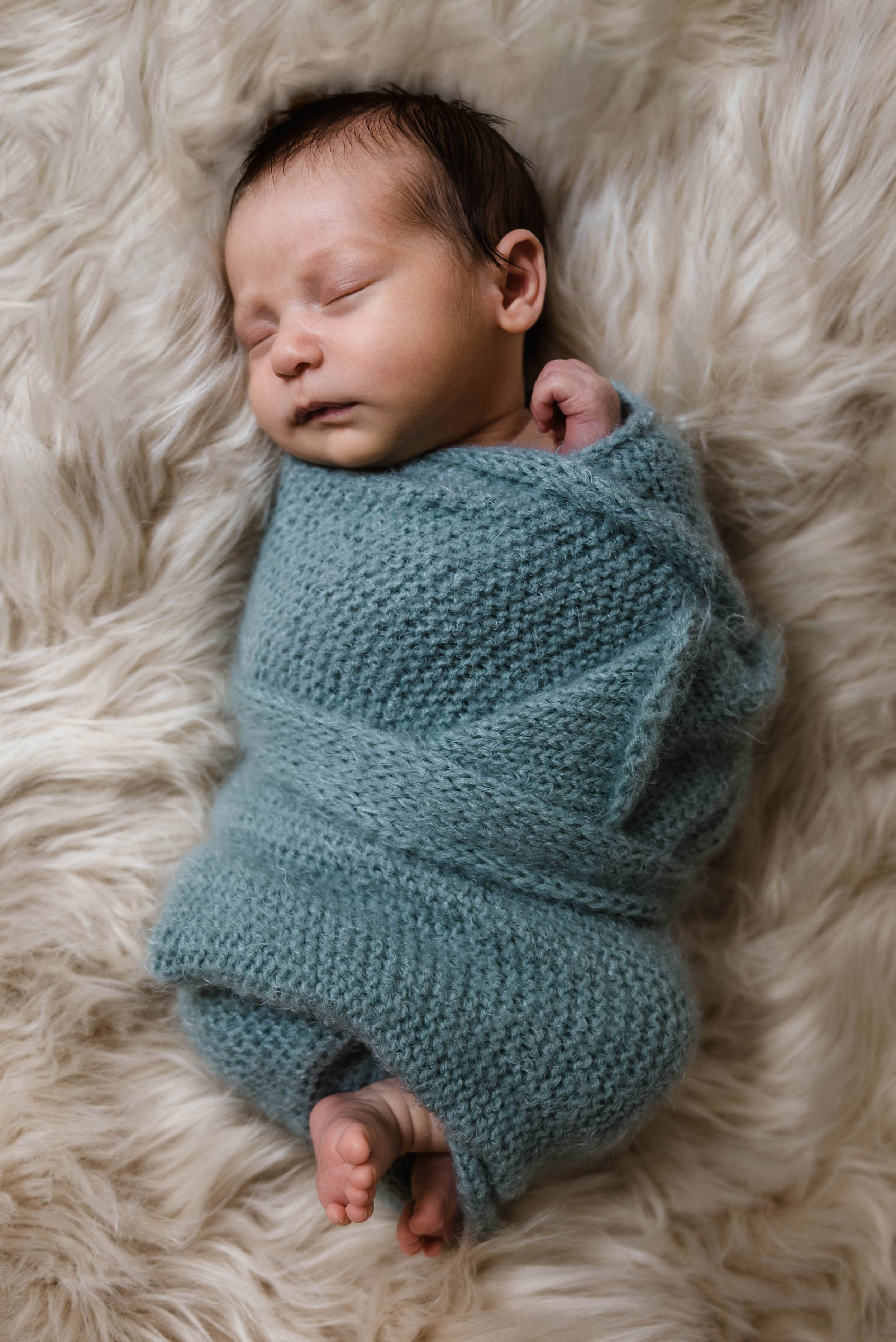 A sleeping newborn baby wrapped in a knitted blue blanket, lying on a fluffy cream-colored surface. Studio newborn portrait, Brownsville PA.