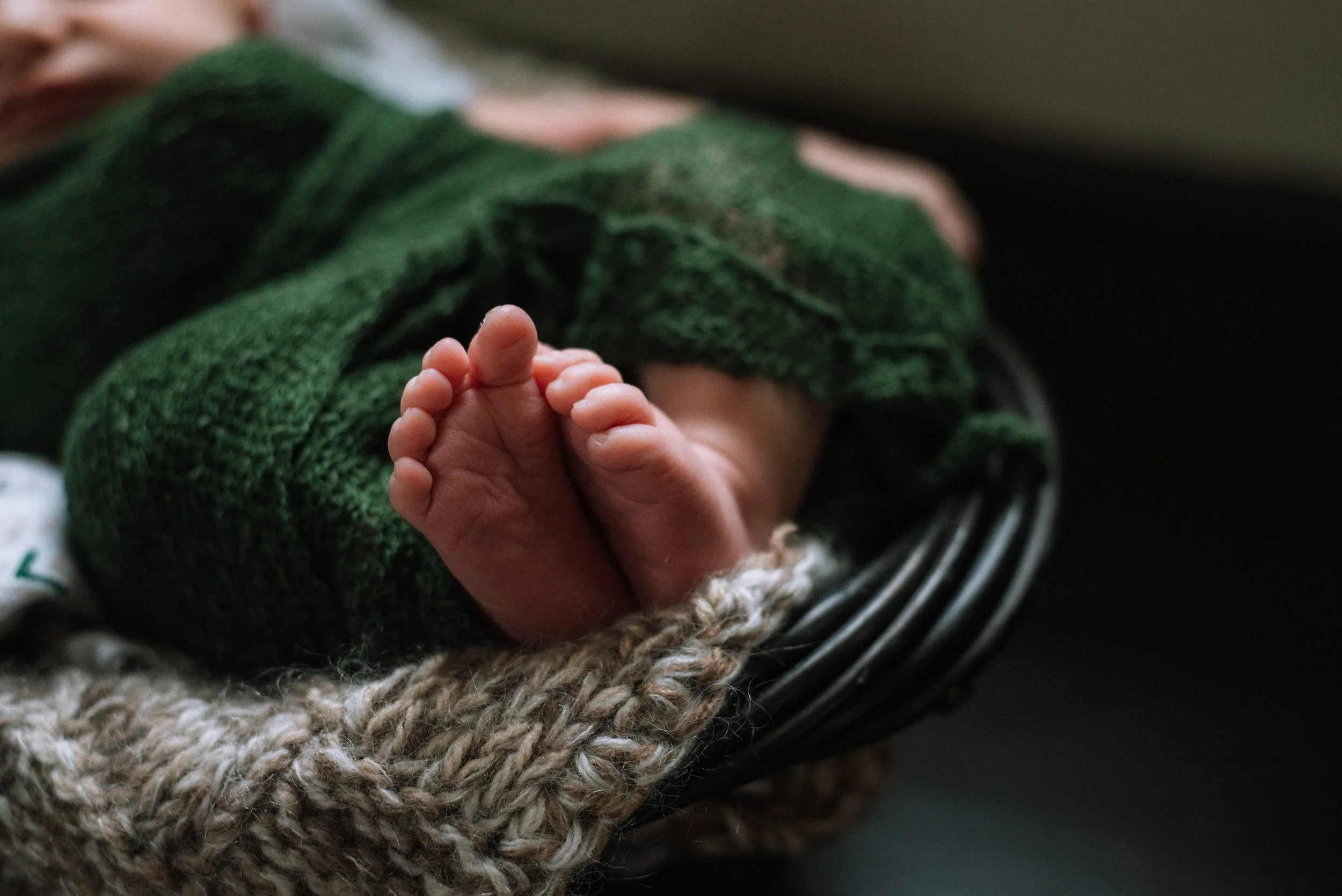 Close-up of a baby's feet with tiny toes, wrapped in a green knitted blanket, resting on a woven beige blanket and a metal surface. Studio newborn portrait, Brownsville PA.
