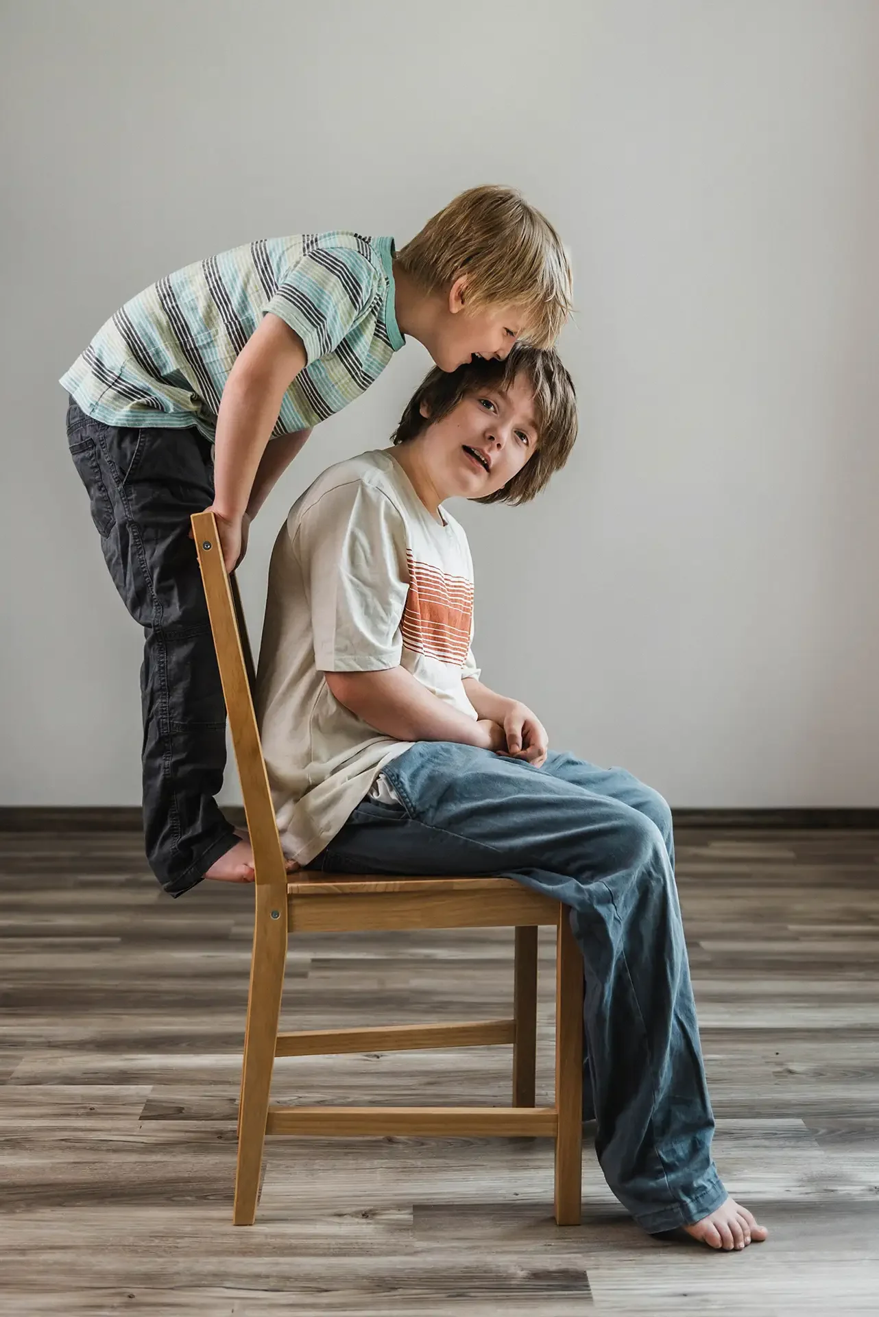 Two young boys in the studio, the older sits in a wooden chair looking at the camera while the younger climbs on the back of the chair to lean into his brother, they are wearing casual clothing