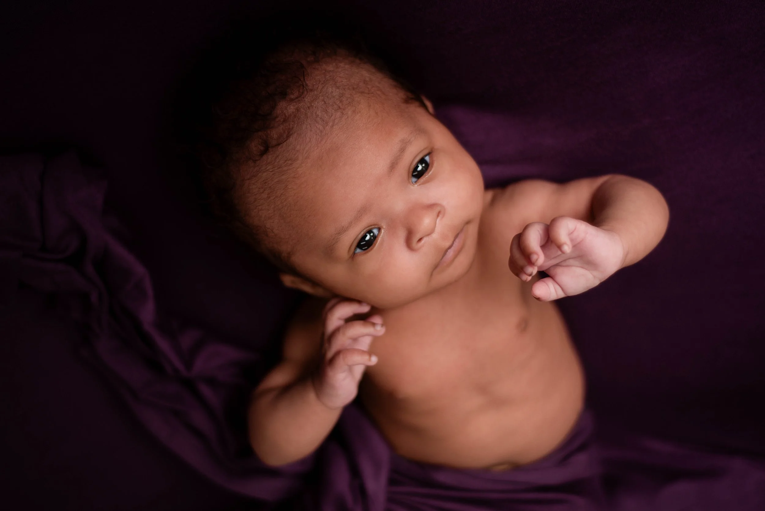 an 8 week old baby with dark skin and curly hair is stretching her arms toward camera, she is wrapped from the waist down in deep plum fabric