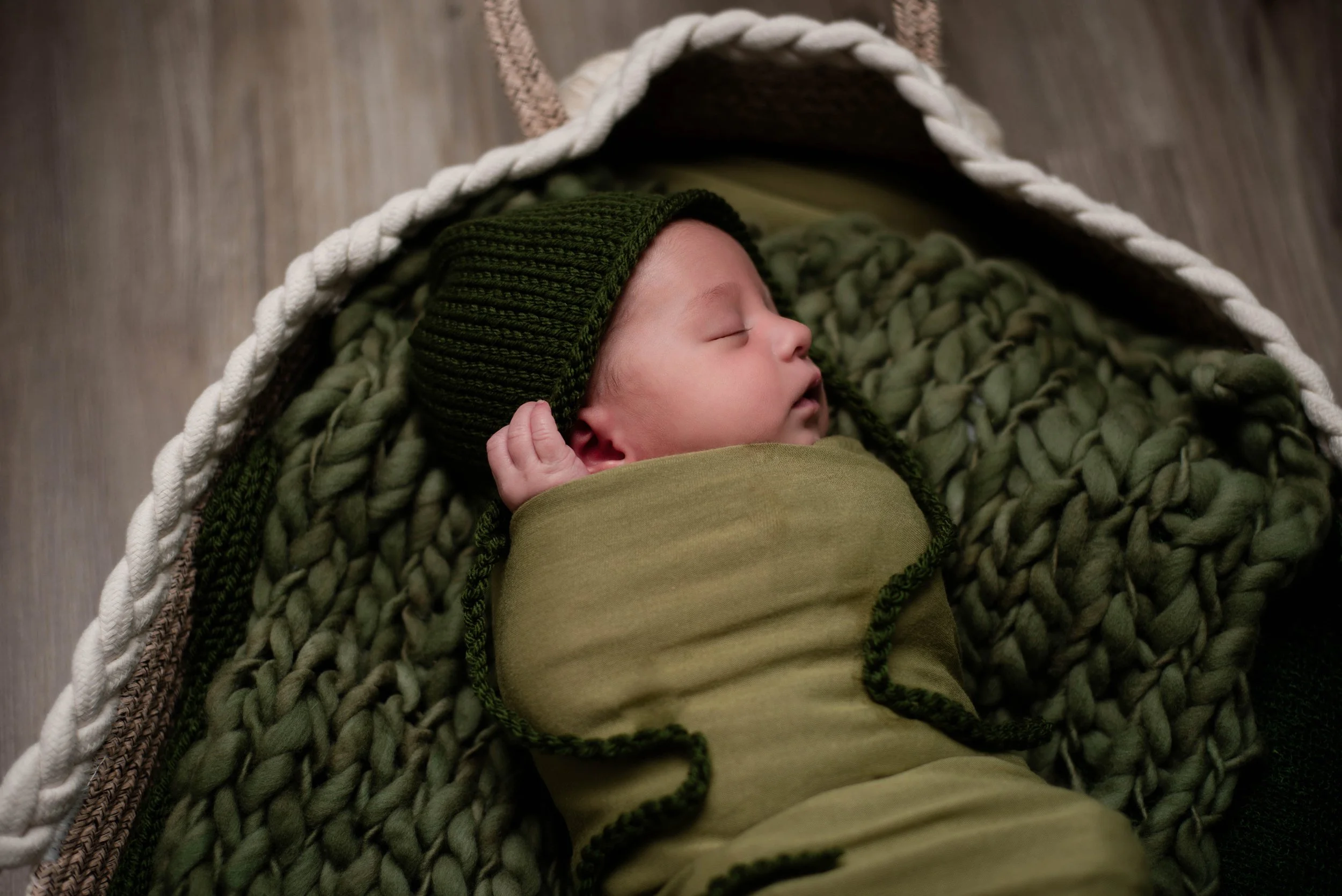A peaceful baby sleeping in a woven basket, wrapped in a green blanket and wearing a green knit hat. Studio newborn portrait, Brownsville PA.