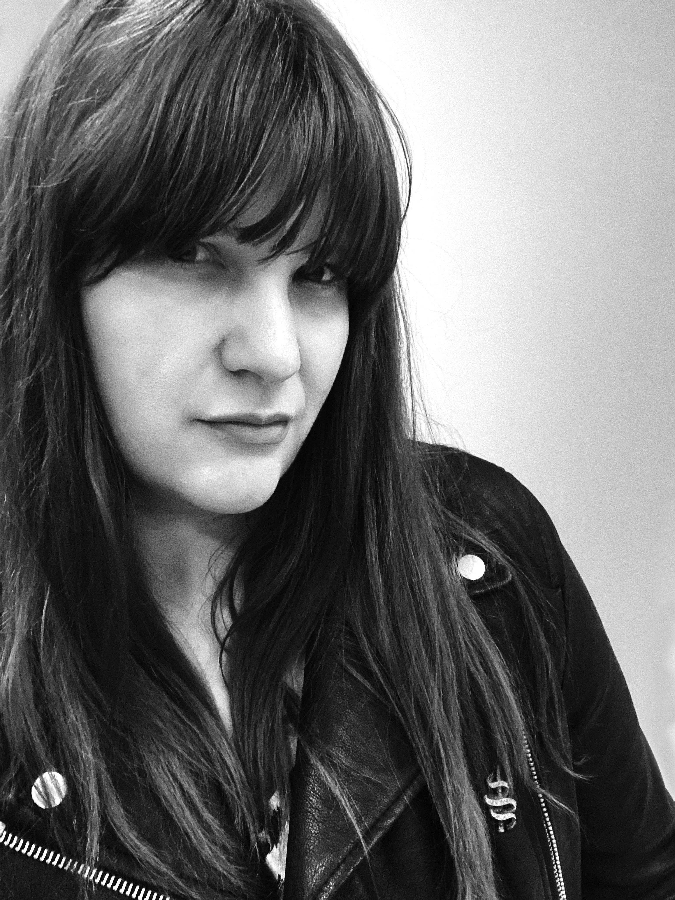Black and white photo of a woman with long, dark hair and bangs, looking slightly to the side, wearing a black leather jacket.