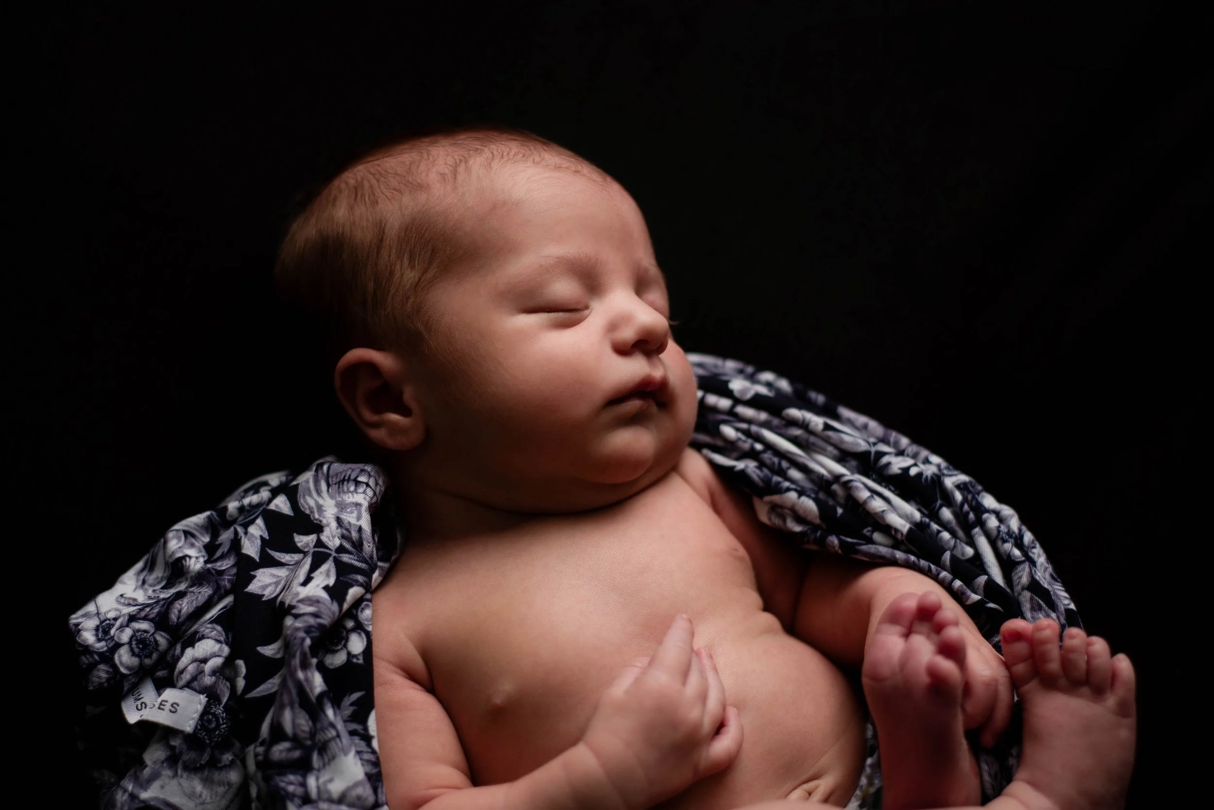 A sleeping baby with closed eyes and a calm expression, lying on a black background, partially wrapped in a black and white patterned cloth with flowers and skulls. Studio newborn portrait, Uniontown PA.