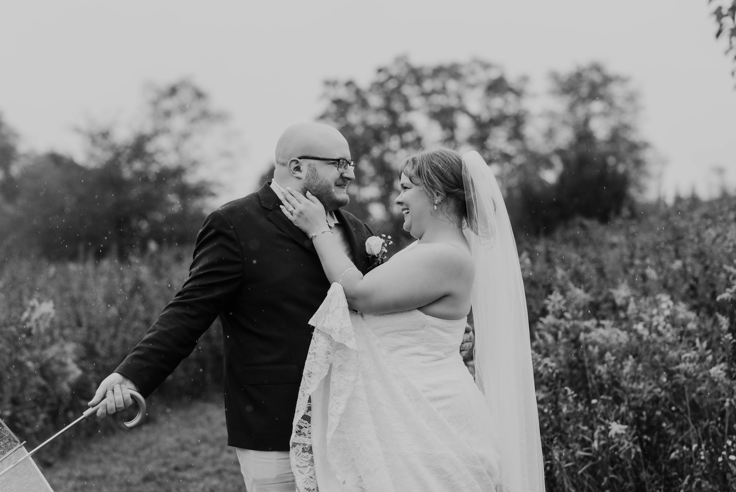 black and white image of a bride and groom standing in a field of flowers in the rain on their wedding day, he is holding an umbrella but not using it