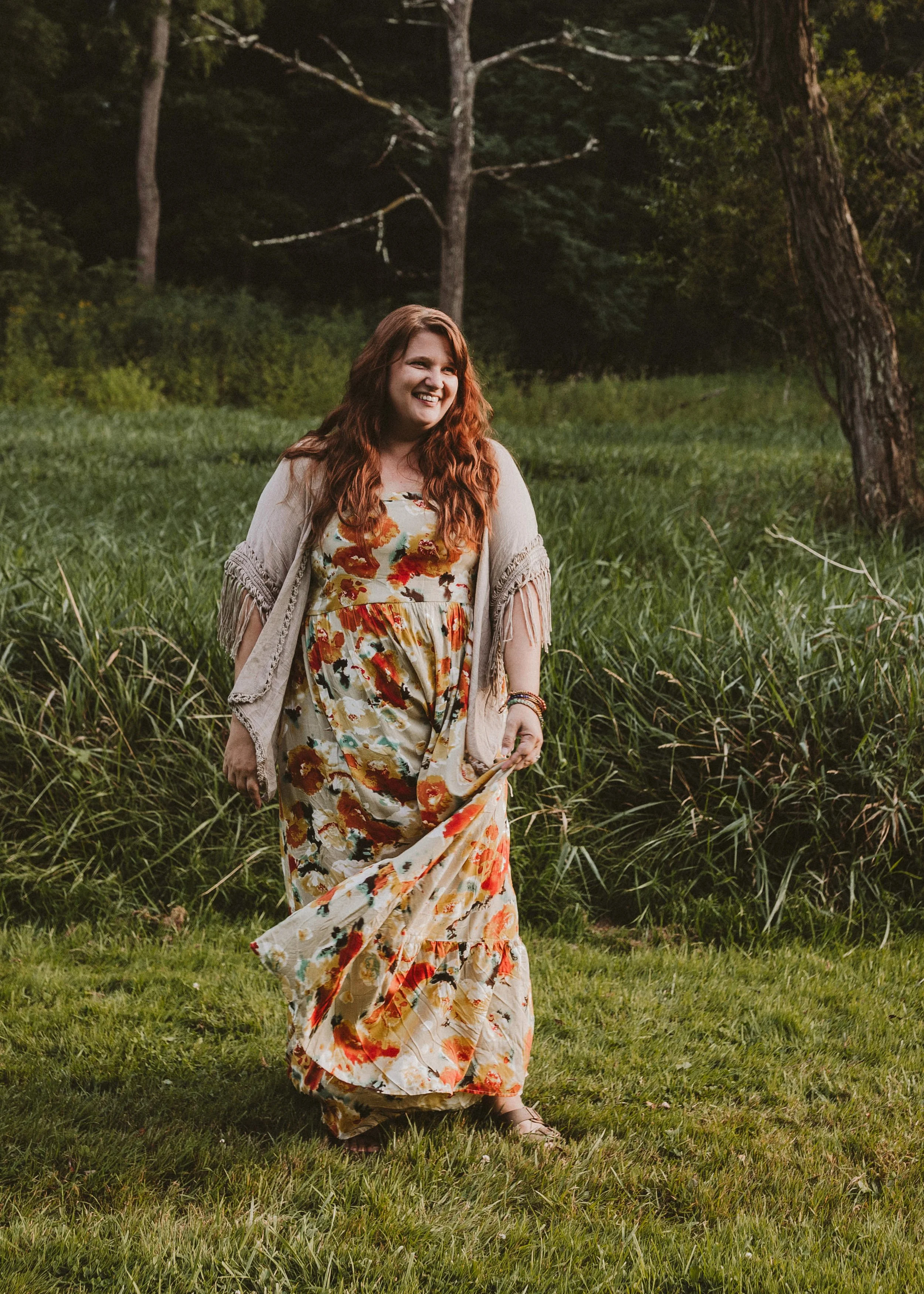 A woman with curly red hair smiling and walking through a grassy field with trees in the background, wearing a floral dress and a light shawl.