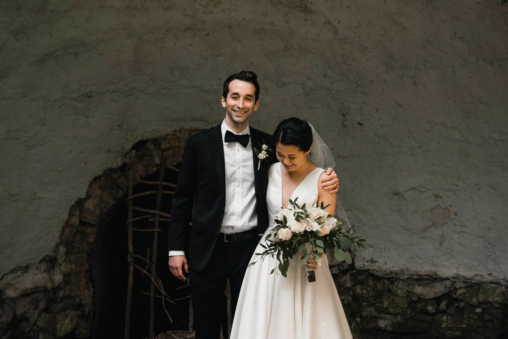 A newlywed couple smiling, standing together in an indoor setting. The man is in a black tuxedo with a bow tie, and the woman is in a white wedding gown holding a bouquet of white and pink flowers. She has a veil and dark hair. The background feature