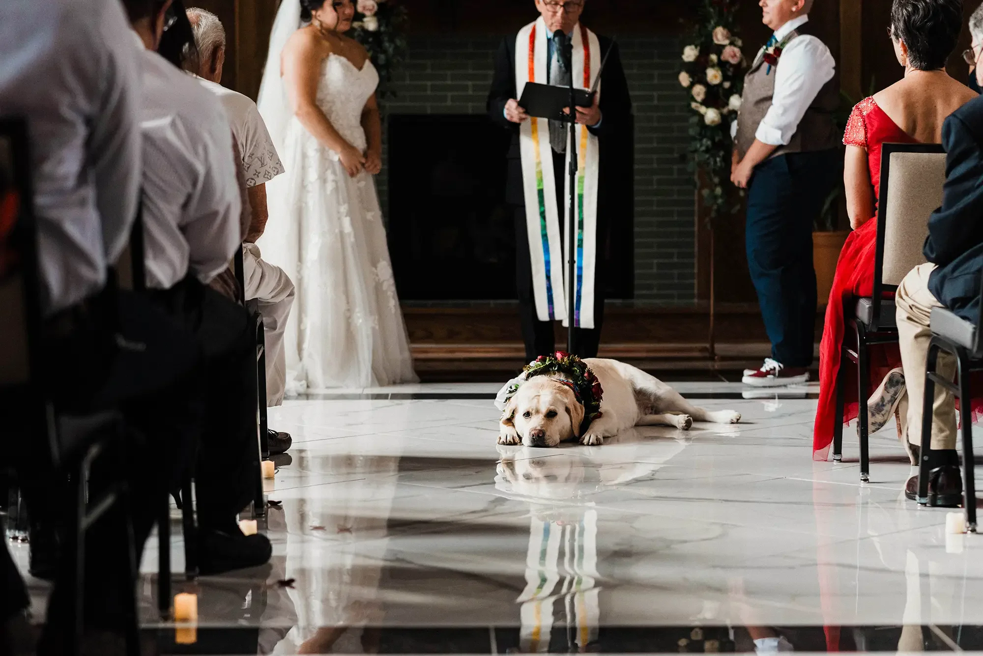 A dog wearing a floral wreath lying down on a shiny white floor during a wedding ceremony, with people standing and sitting around, and a bride in a white dress in the background. Hotel Morgan, LGBT wedding, West Virginia.