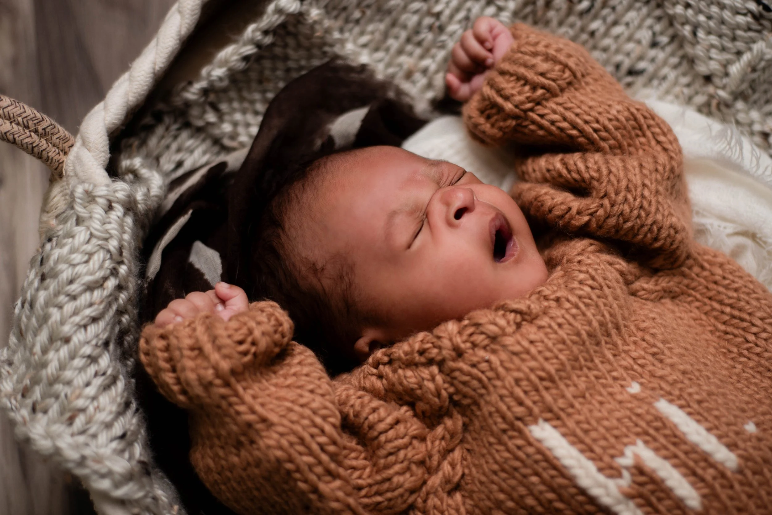 A newborn baby wearing a brown knitted sweater yawns while lying in a textured cream-colored basket. Studio newborn portrait, Brownsville PA.