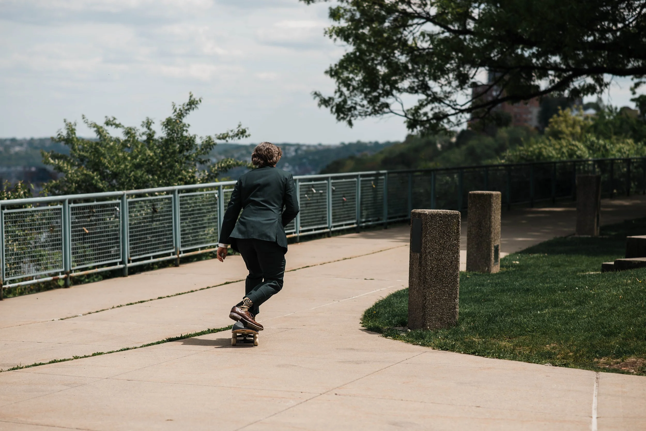 Groom in a suit and dress shoes rides a skateboard away from camera at West End Overlook elopement