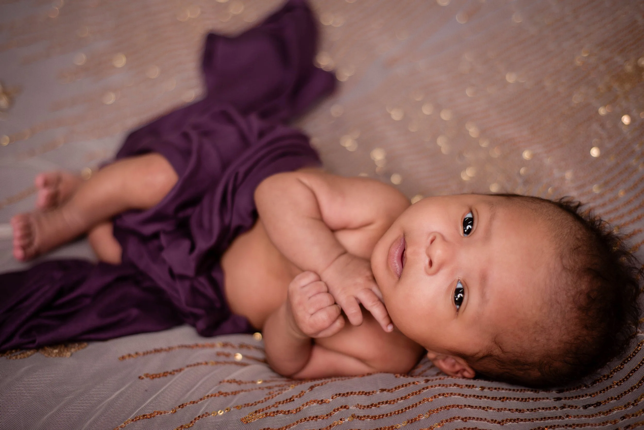 A baby lying on a fabric with gold and pink sequins, wearing only purple fabric draped around them, with arms crossed and looking at the camera. Studio newborn portrait, Uniontown PA.