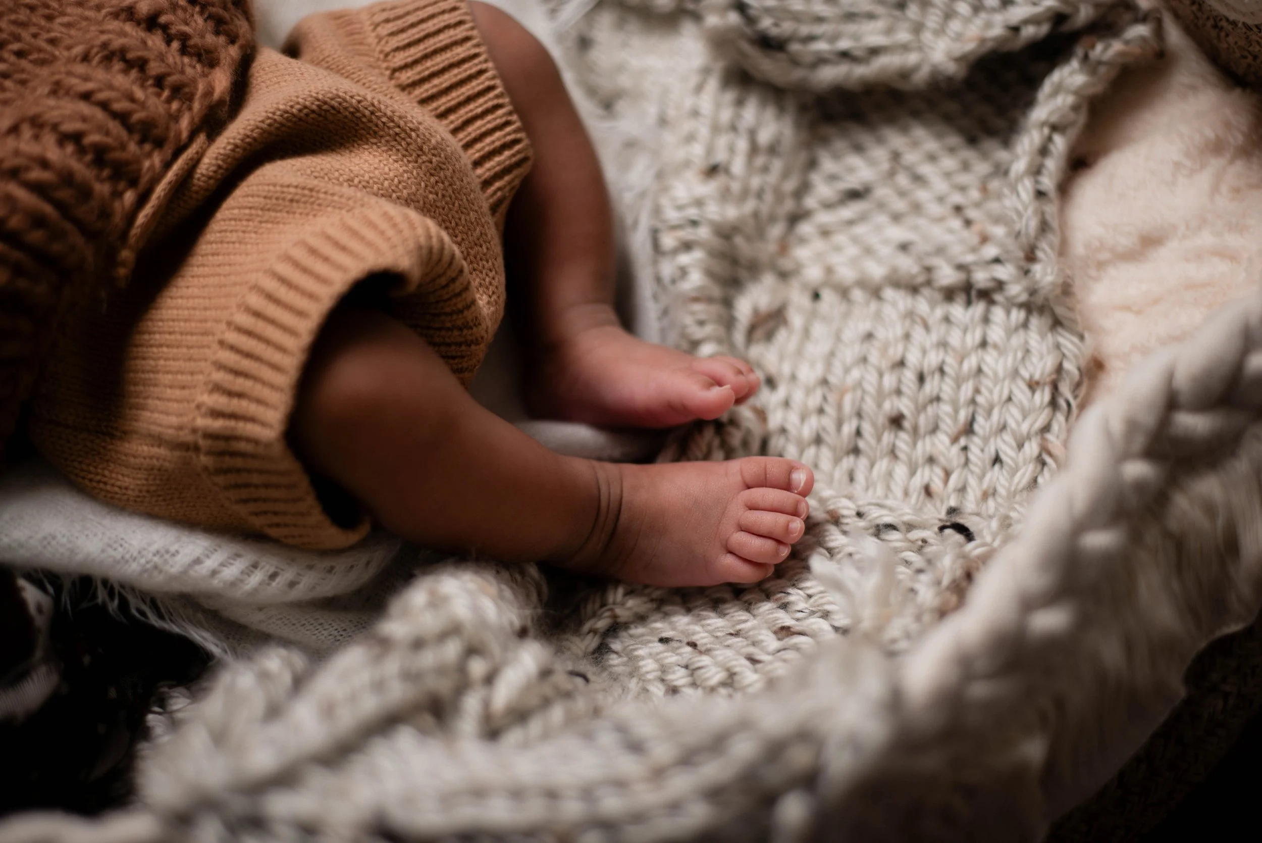 Close-up of a baby's legs and feet lying on a chunky knit blanket, wearing a brown sweater and orange knit pants. Studio newborn portrait, Brownsville PA.