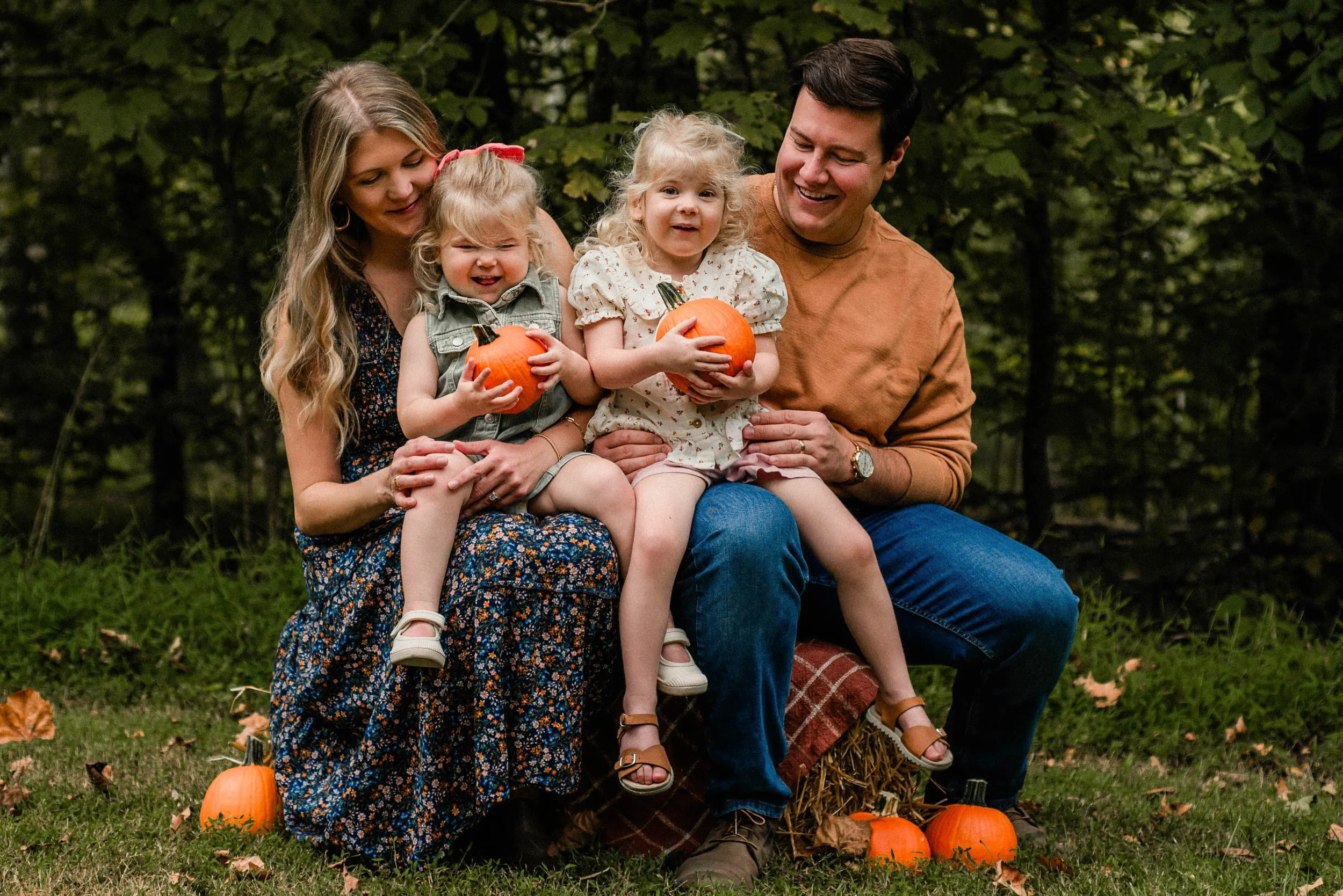 a whole family, dad, mom, and two young girls, sit on a hay bale at cedar creek park for their family mini sessions. they are holding pumpkins.