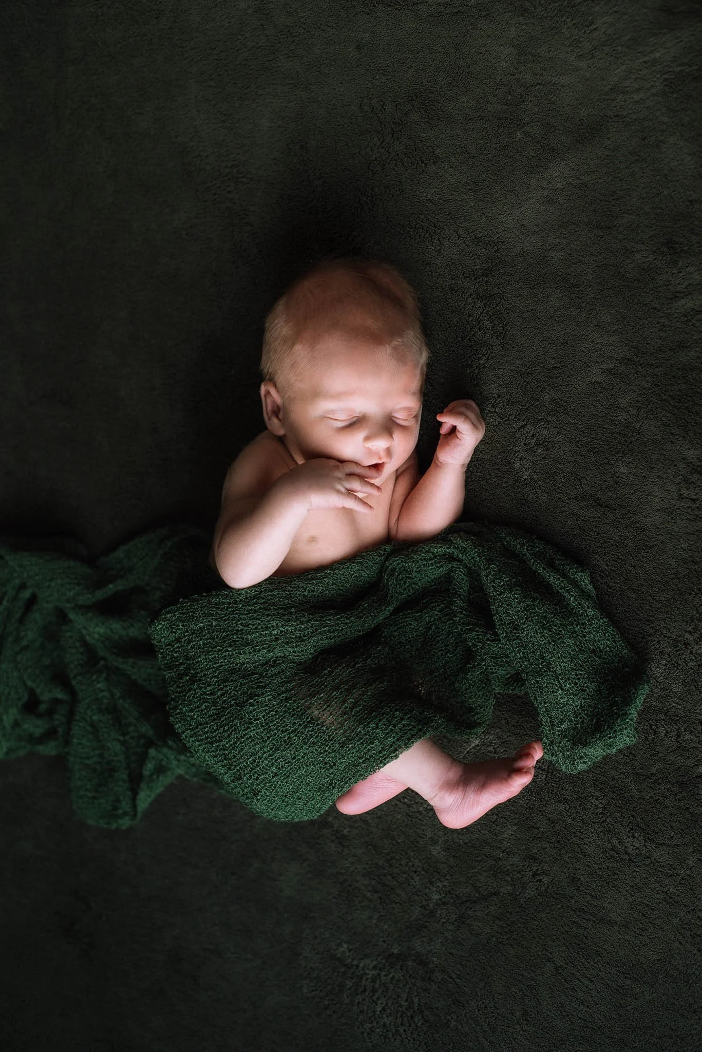 A newborn baby with light reddish hair sleeping on a dark textured surface, partially covered with a dark green towel, with one hand near the face and the other arm bent. Studio newborn portrait, Brownsville PA.