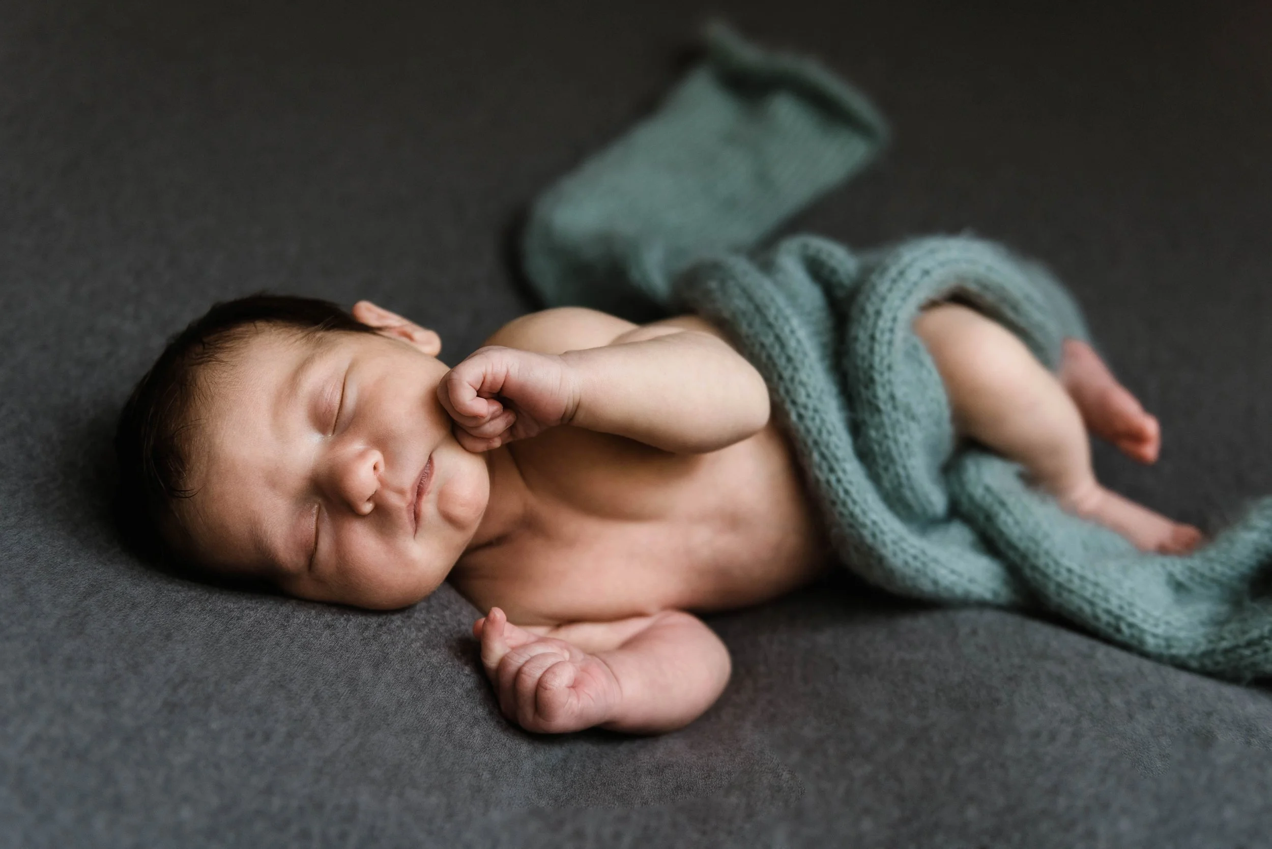 A newborn baby sleeping on a dark gray surface, partially covered with a knitted blue blanket, with a peaceful expression and tiny fists near his face. Studio newborn portrait, Brownsville PA.
