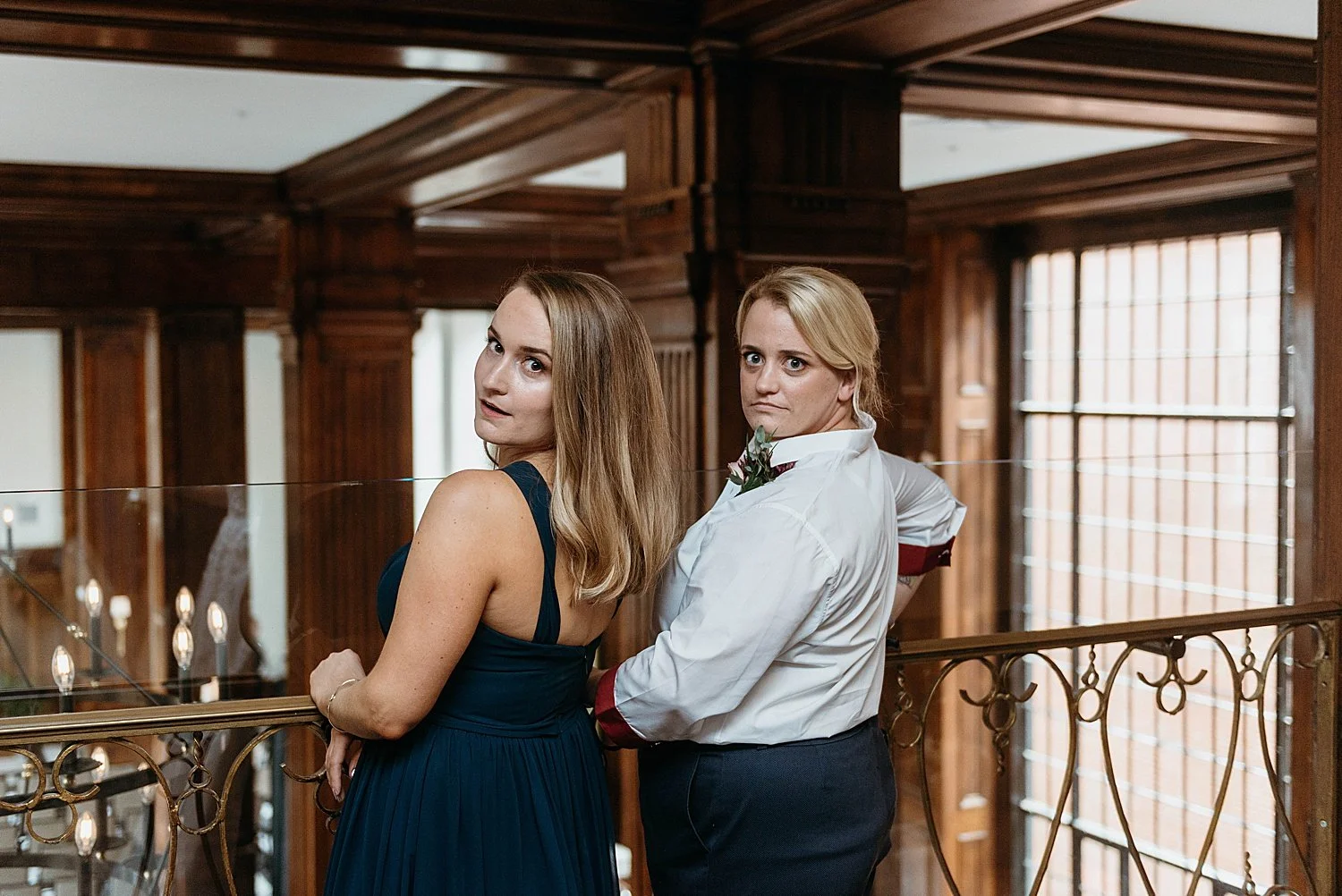 Two women lean back from railing with silly surprised faces. One is wearing a blue dress, the other a white button down shirt.