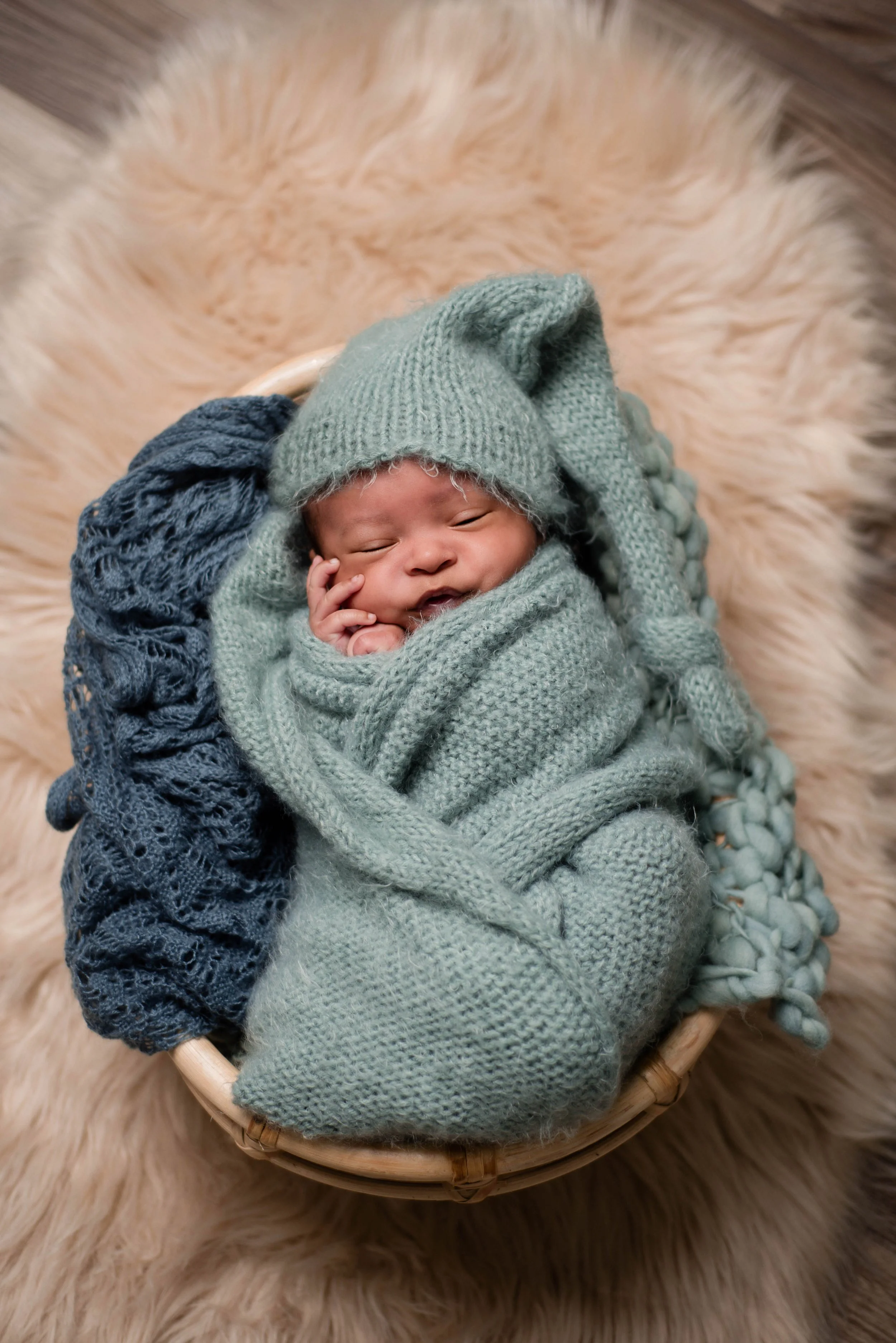 A newborn baby wrapped in a cozy blue knitted blanket and hat, lying in a woven basket on a soft, fluffy beige rug. Studio newborn portrait, Brownsville PA.