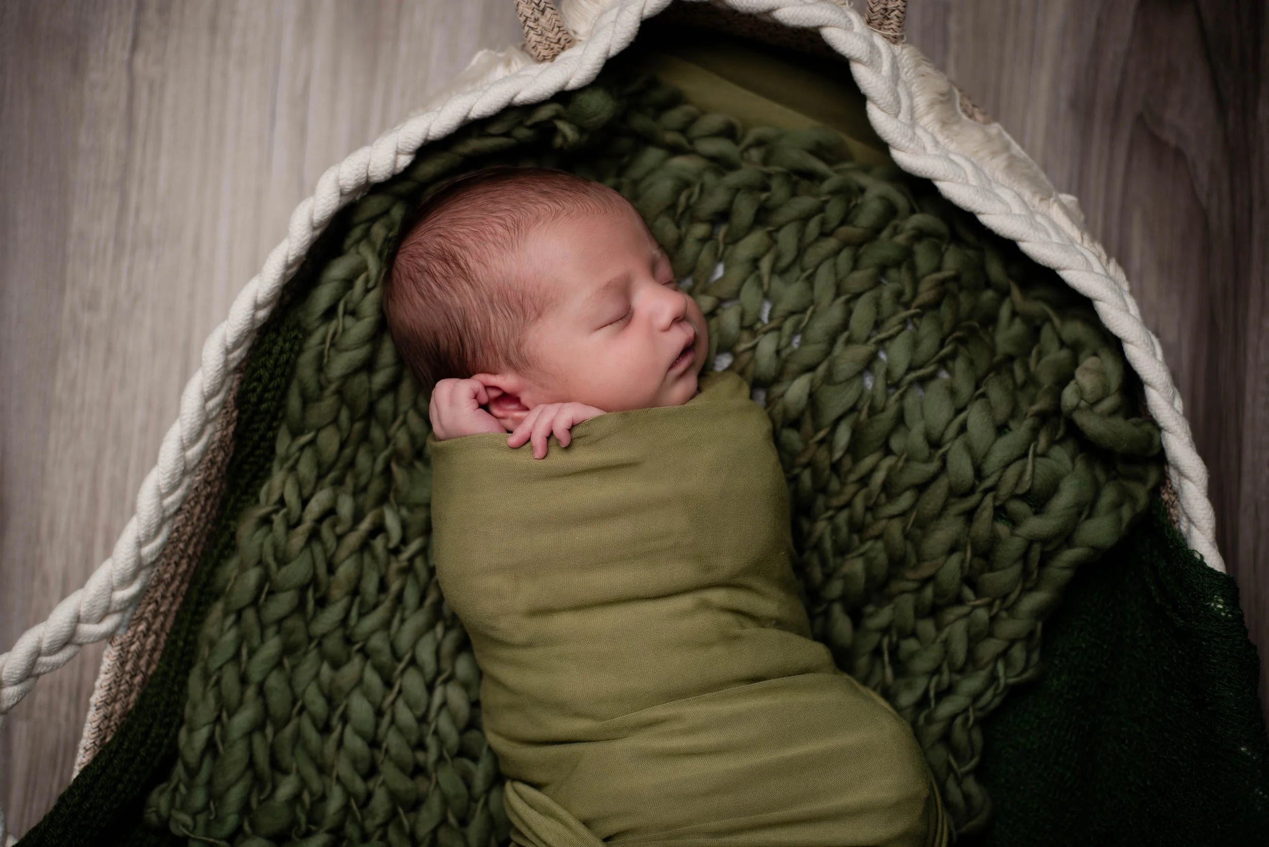 A newborn baby sleeping peacefully, swaddled in green cloth, lying on a textured green blanket inside a woven basket with a rounded edge, on a wooden floor. Studio newborn portrait, Brownsville PA.