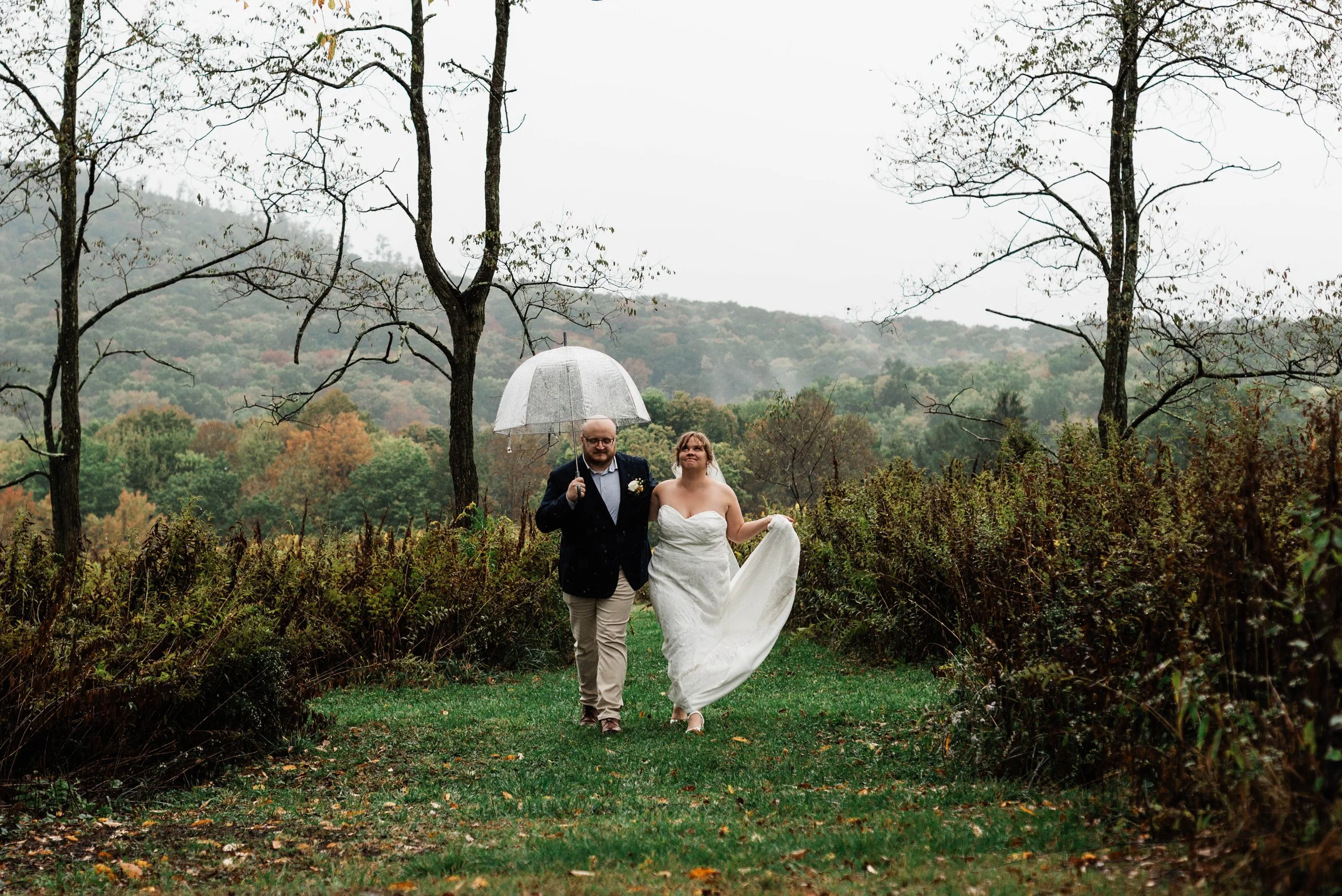 a man in a dark suit jacket and khaki pants holds an umbrella over his head, not realizing he is leaving his bride in her long strapless gown out in the rain