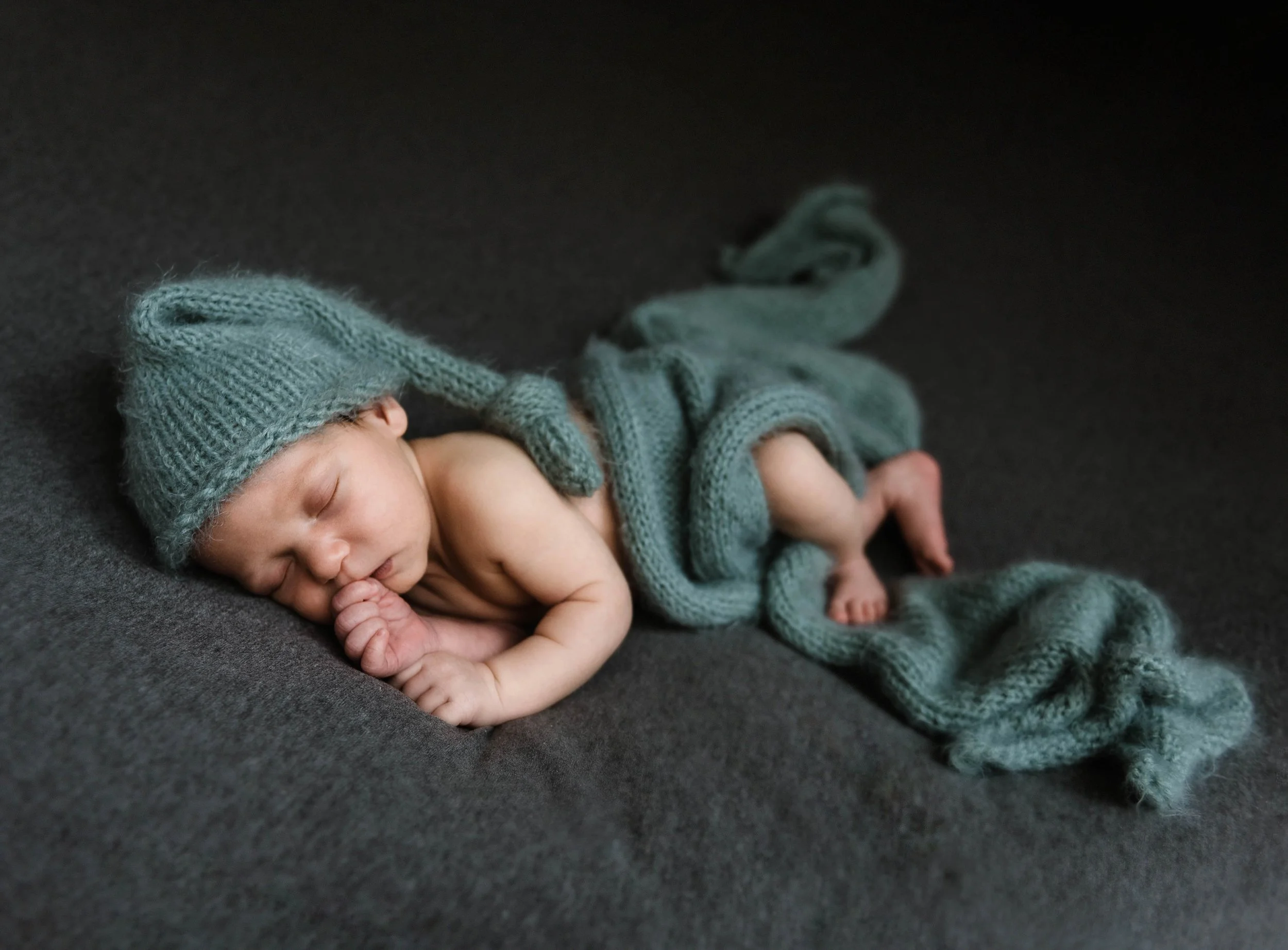 A newborn baby sleeping on a dark surface, wearing a knitted hat and wrap. Studio newborn portrait, Brownsville PA.