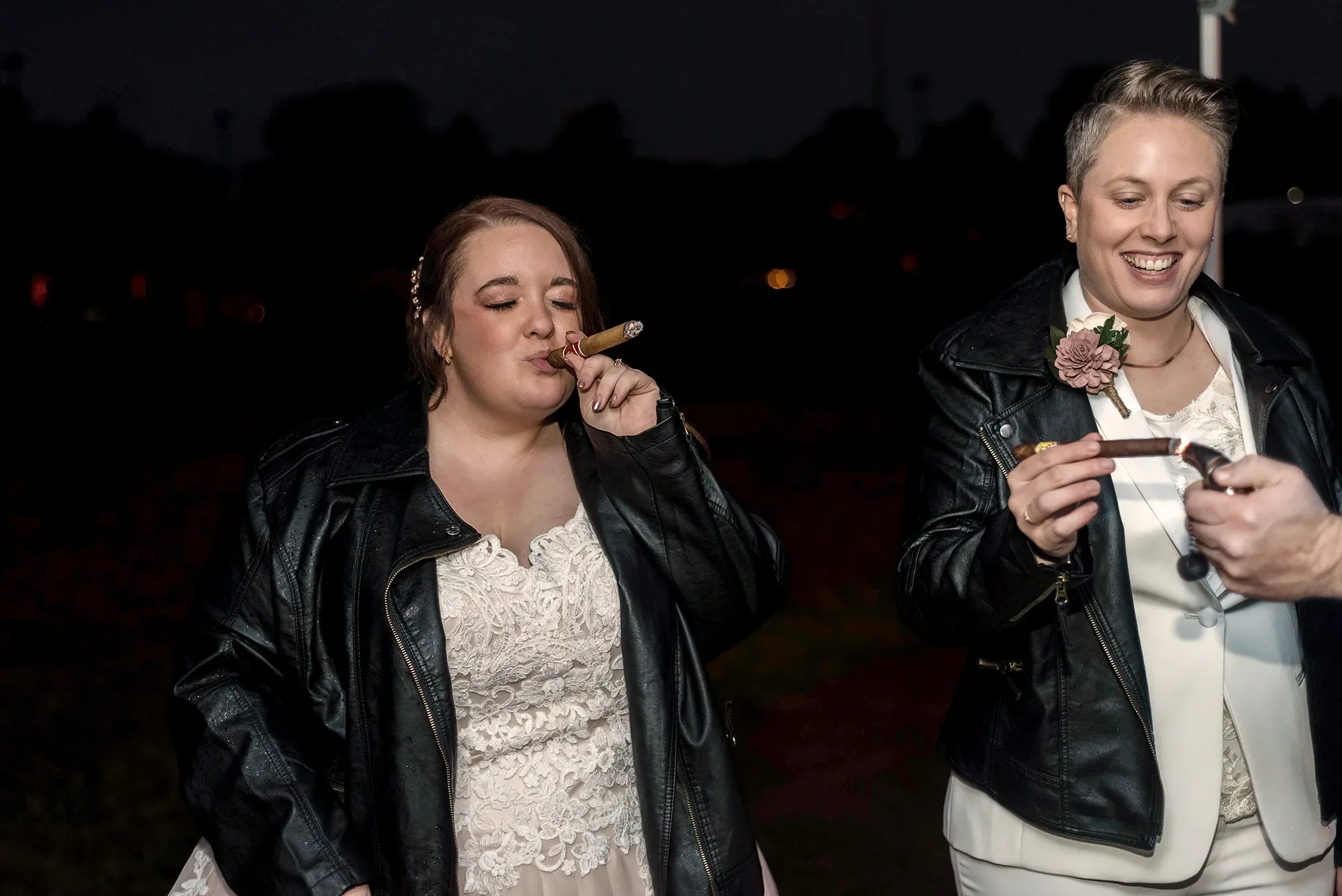 Two women wearing leather jackets and white dresses, one smoking a cigar at night. Queer elopement at the Pump House in Pittsburgh, PA.
