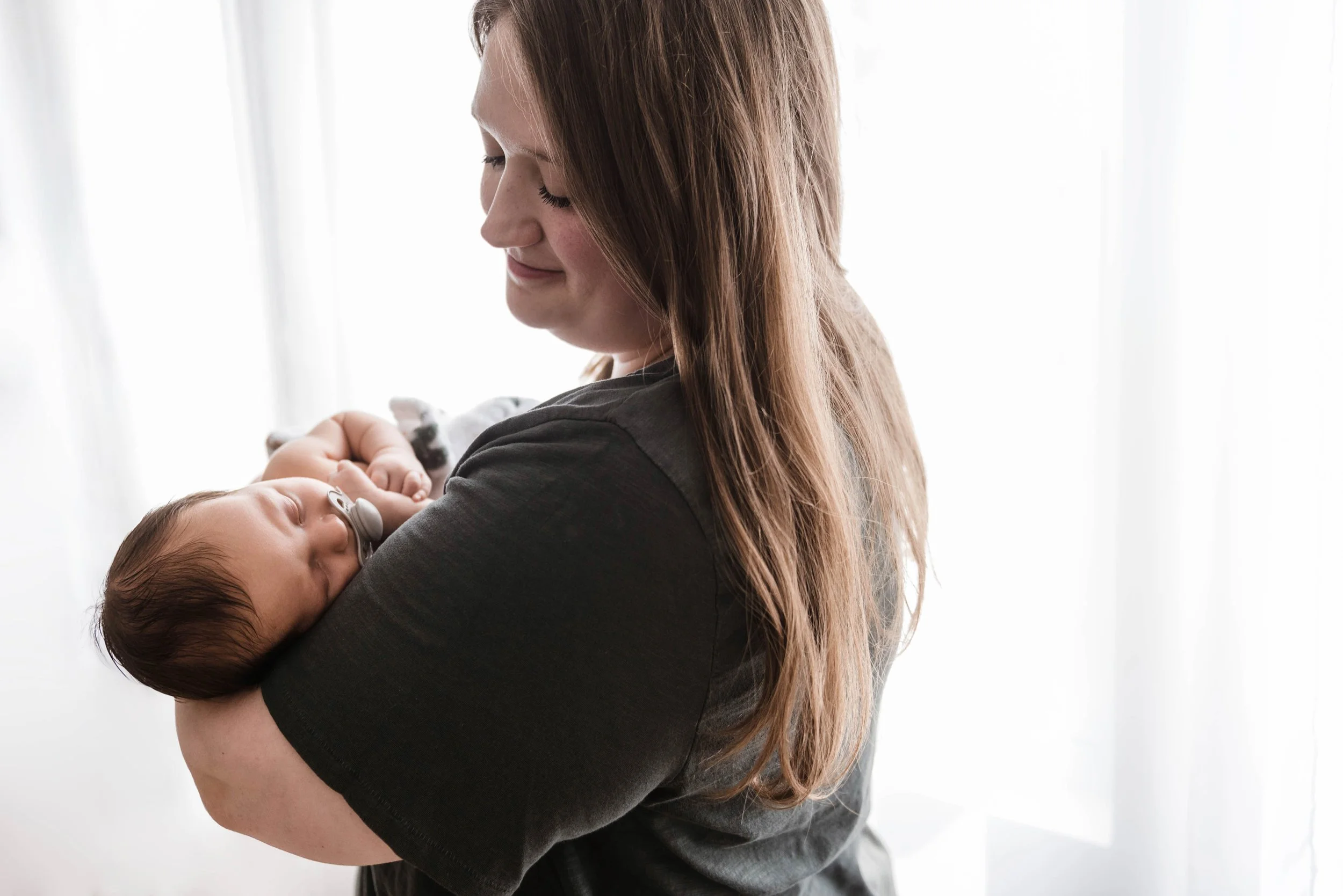 A woman with long hair holding a sleeping baby with a pacifier in a well-lit room. Studio newborn portrait, Brownsville PA.