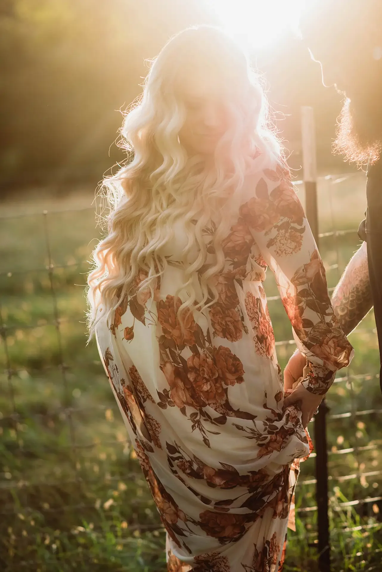 a pregnant woman in a long floral dress stands in front of the sunset, there is a flare in the camera and she looks glowy, she holds hands with her husband who is just in frame