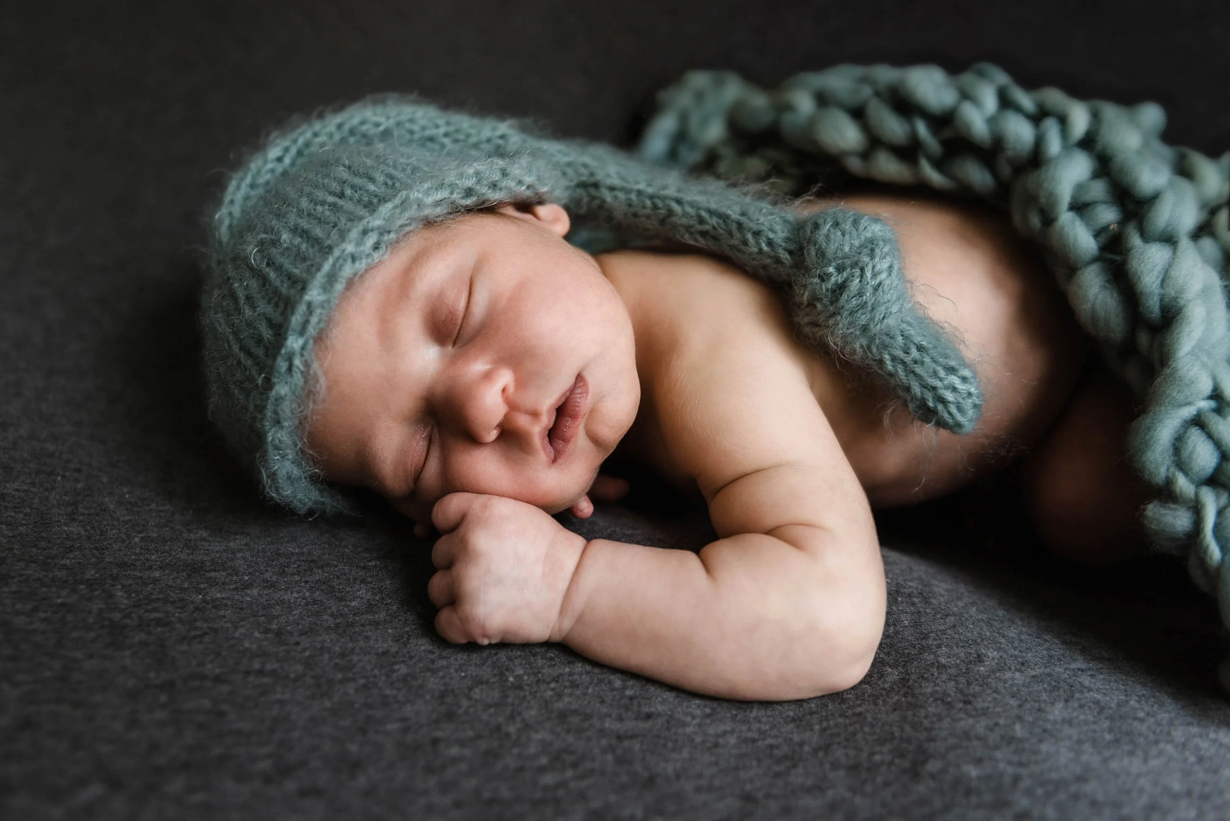 A sleeping newborn baby wearing a knitted blue hat and blanket, lying on a dark surface. Studio newborn portrait, Brownsville PA.