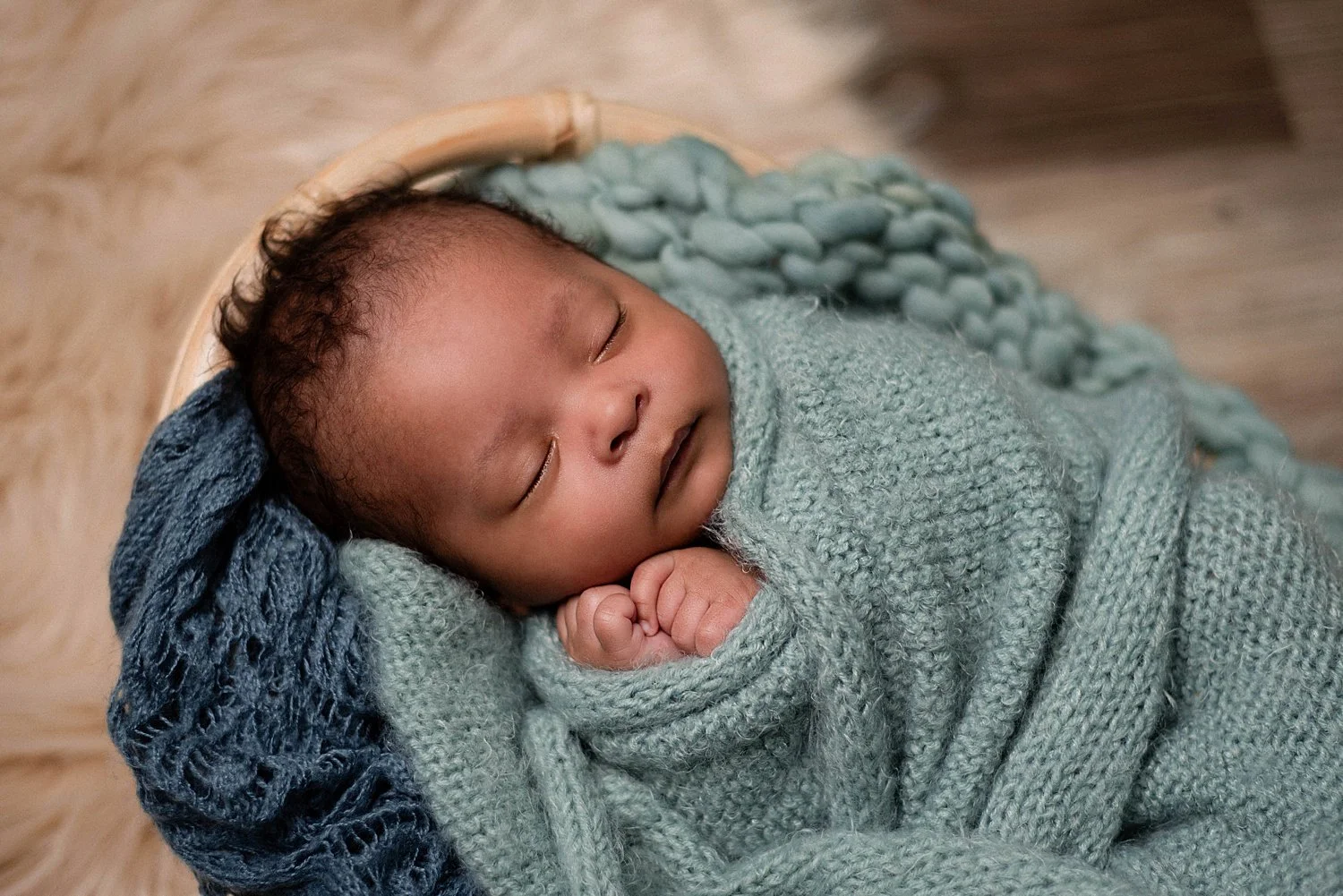 sleeping newborn, dark skinned with curly hair, hands are curled up by face, blue knit swaddle