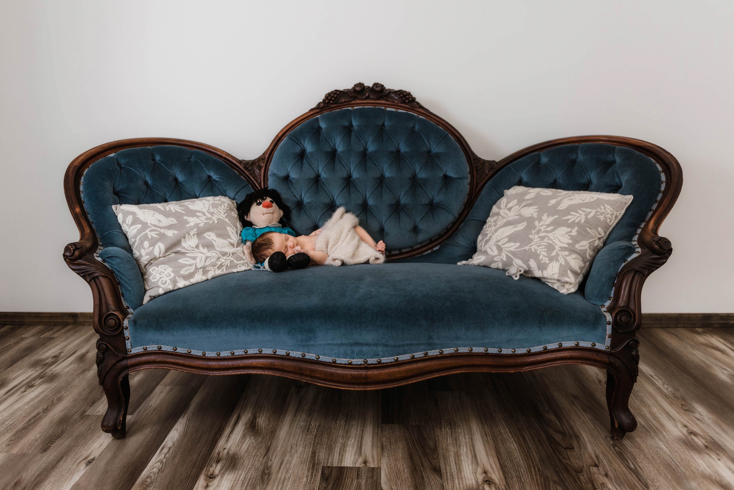 A small child sleeping on a vintage blue velvet sofa with wooden trim, flanked by two patterned gray pillows, with a plush toy from The Big Blue Couch lying beside him. Studio newborn portrait, Brownsville PA.