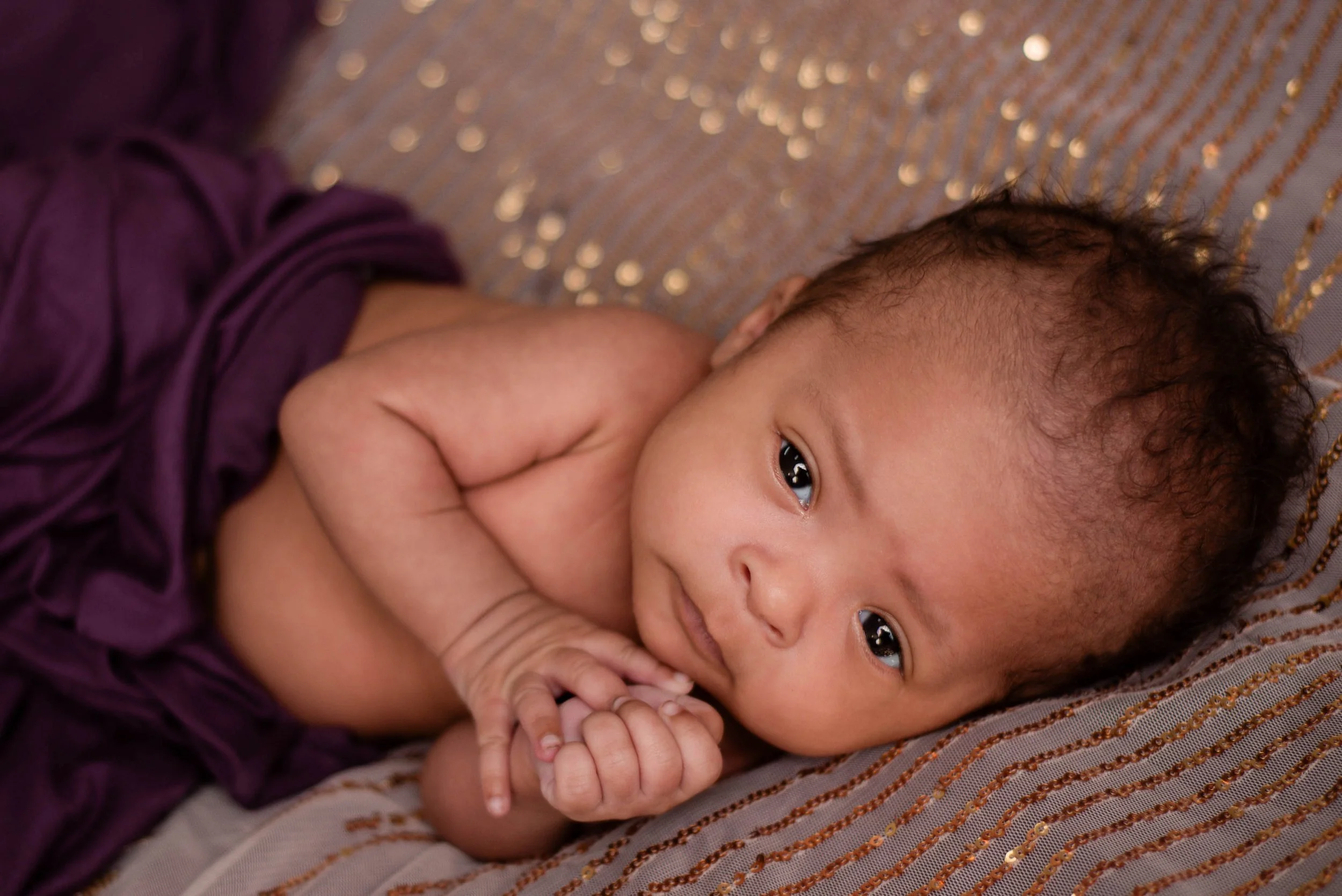 A baby lying on a bed, looking at the camera, with one hand near their mouth and wearing a purple cloth. Studio newborn portrait, Brownsville PA.