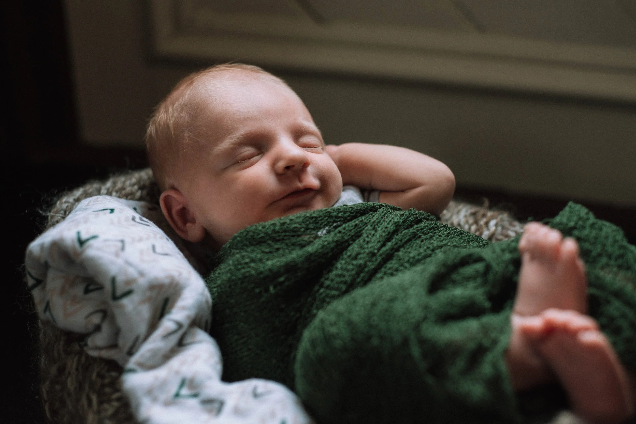 A sleeping baby lying on a cushioned surface, wrapped in a green blanket, with a white pillow behind his head, and a white shirt with colorful patterns. Studio newborn portrait, Brownsville PA.