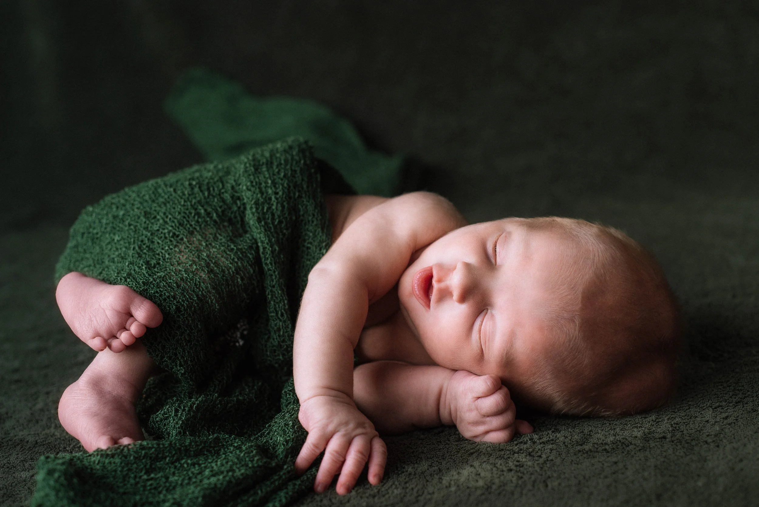 A sleeping newborn baby wrapped in a green blanket on a dark green surface. Studio newborn portrait, Brownsville PA.