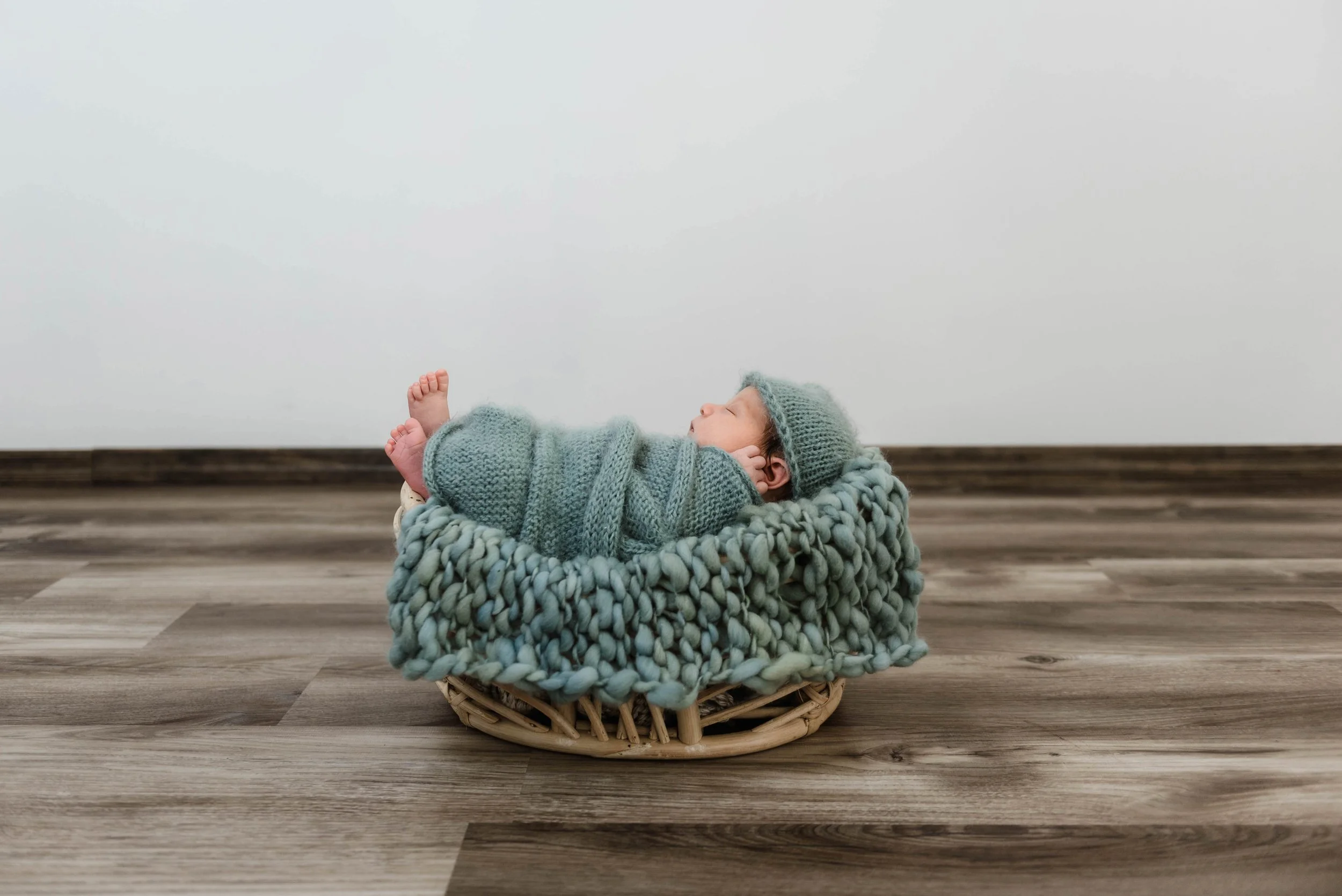 A baby dressed in knitted blue clothes, including a hat, lying on their side in a woven basket with a chunky knit blanket, on a wooden floor against a plain white wall. Studio newborn portrait, Brownsville PA.