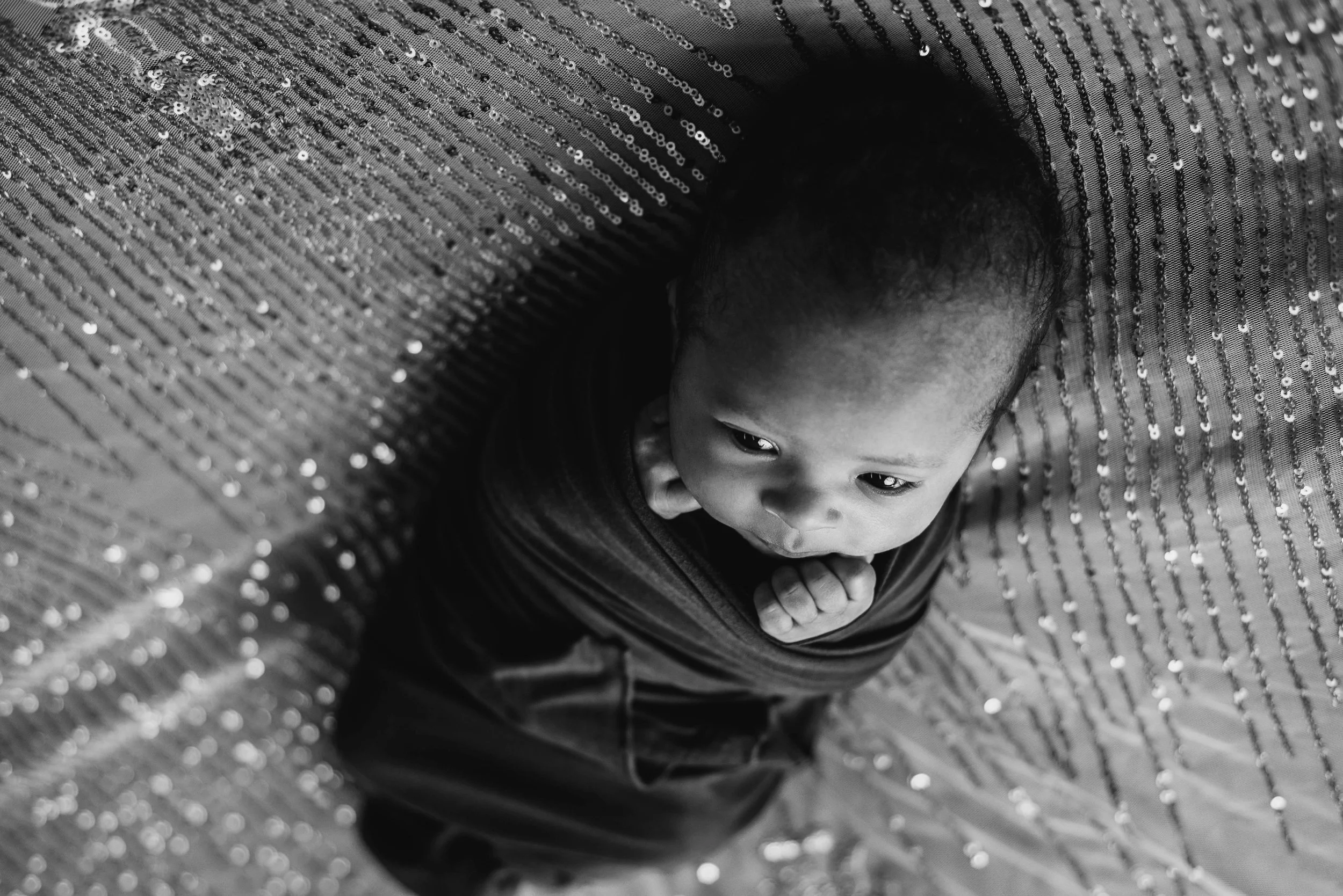 A young child peeking out from under a shiny, sequined fabric or curtain, with a curious expression on their face. Studio newborn portrait, Brownsville PA.