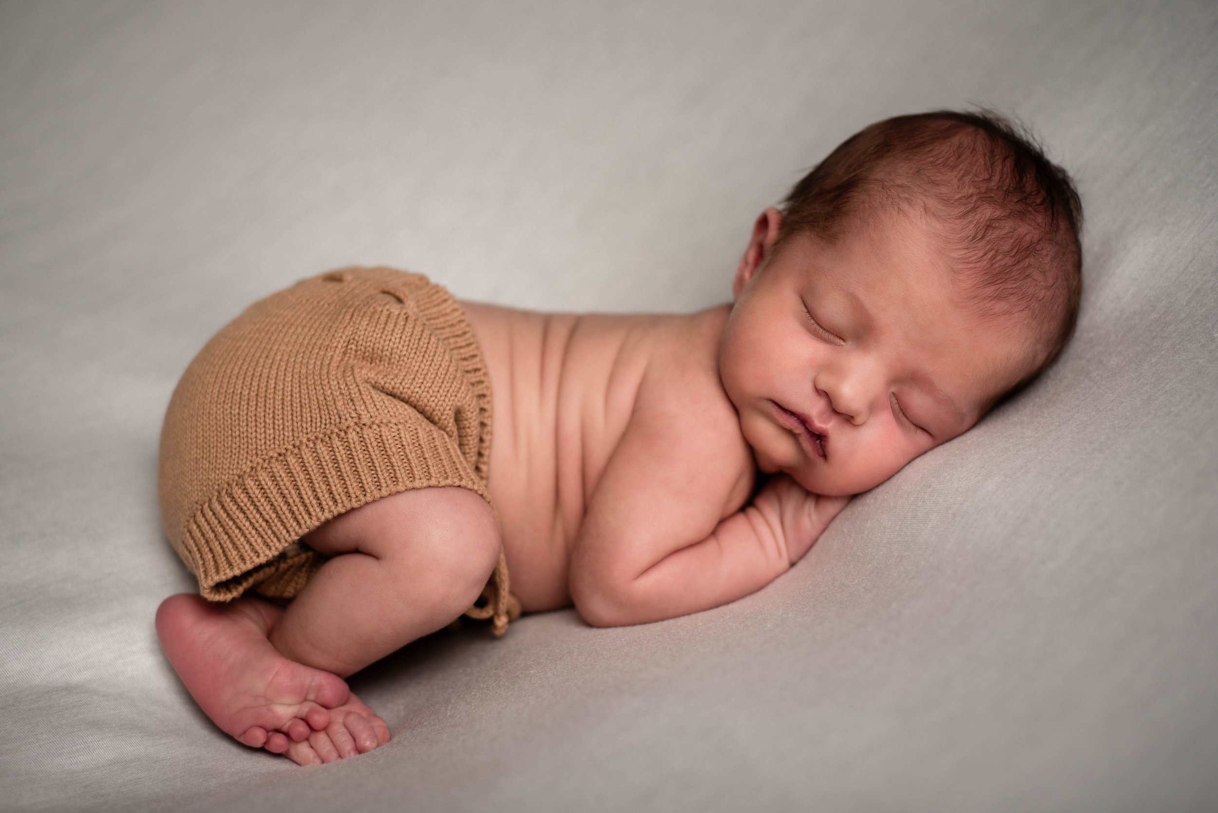 Close-up of a sleeping newborn baby lying on its side on a light gray surface, wearing tan knit shorts. Studio newborn portrait, Brownsville PA.