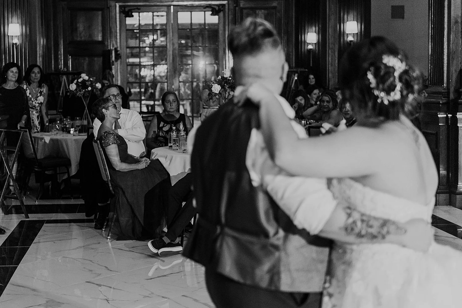 in the foreground, you can see the backs of the bride and groom. they are out of focus, arms around each other. beyond them you can see guests seated at their reception.