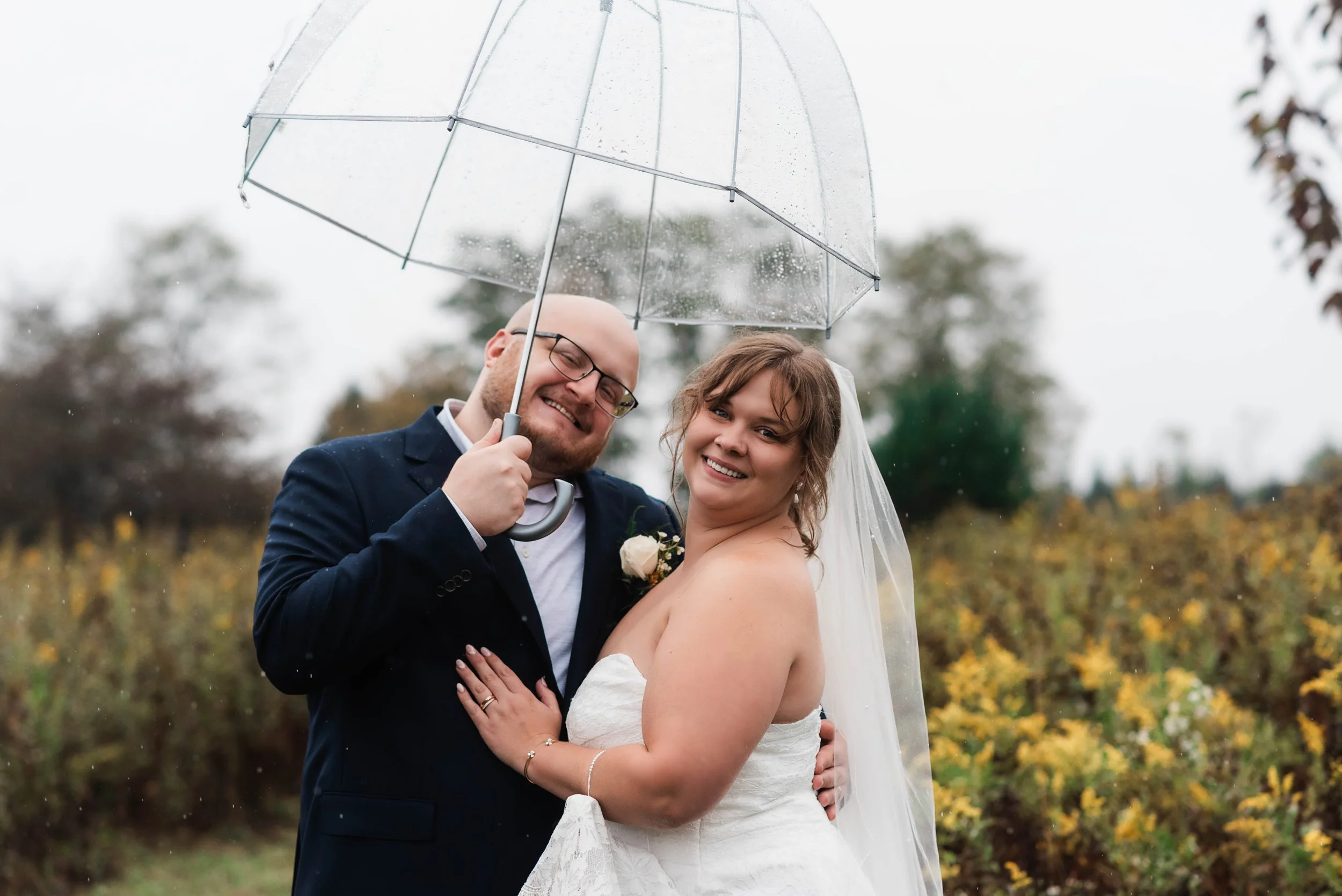 Couple smile at camera on their wedding day, they are holding a clear umbrella because it is raining, behind them are yellow flowers and greenery