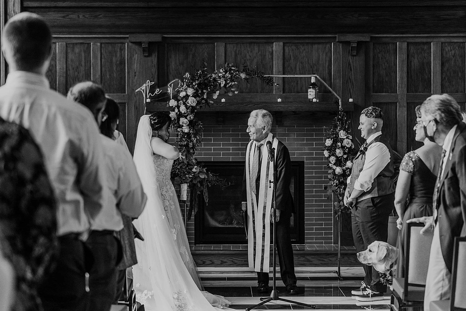 black and white photo of the bride and groom with their officiant. behind them is a fireplace with a floral and metal arch, and a neon sign with their last name. photographed at hotel morgan.