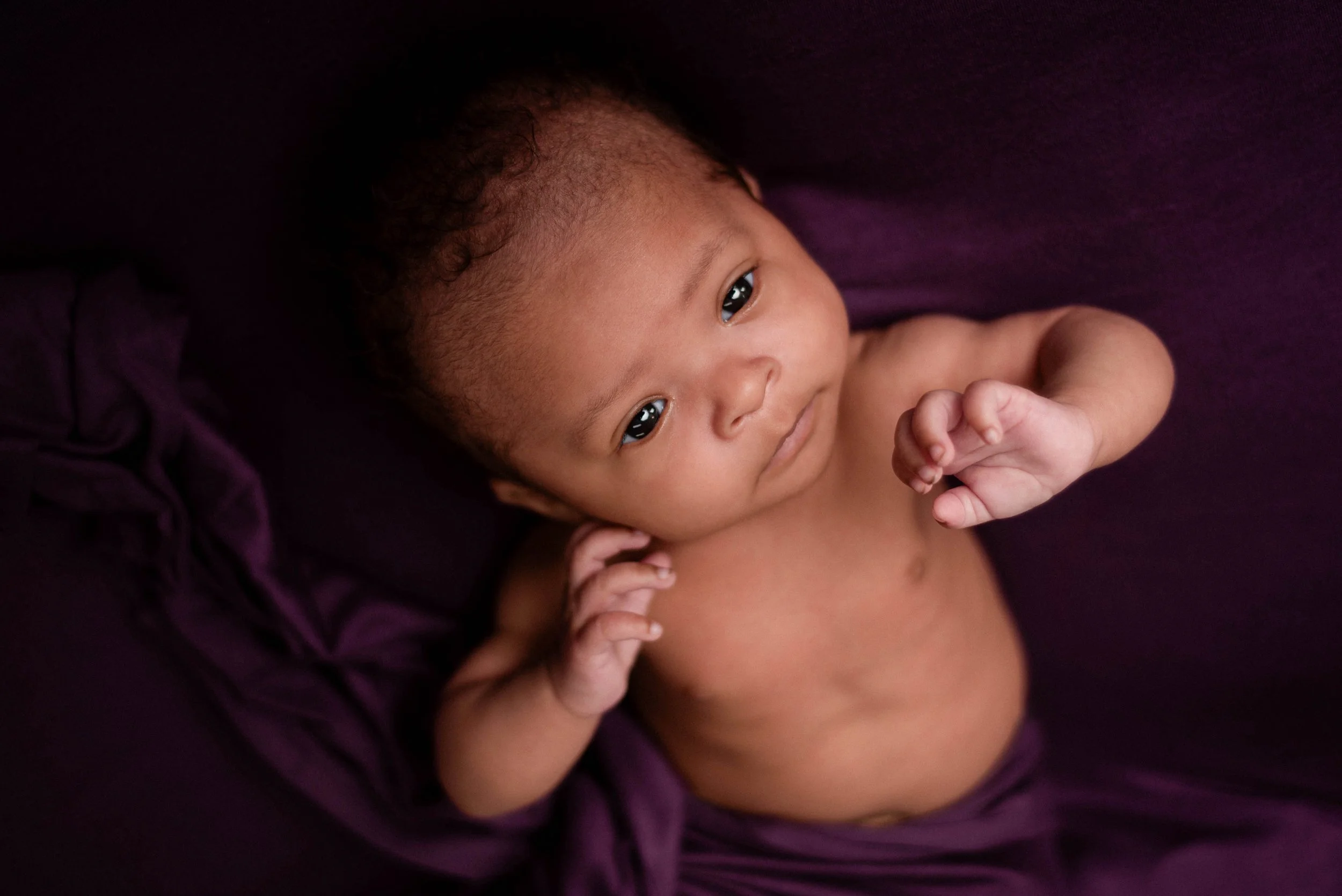 black newborn stretches her arms toward camera, her eyes are open and curious. background is a deep plum fabric.