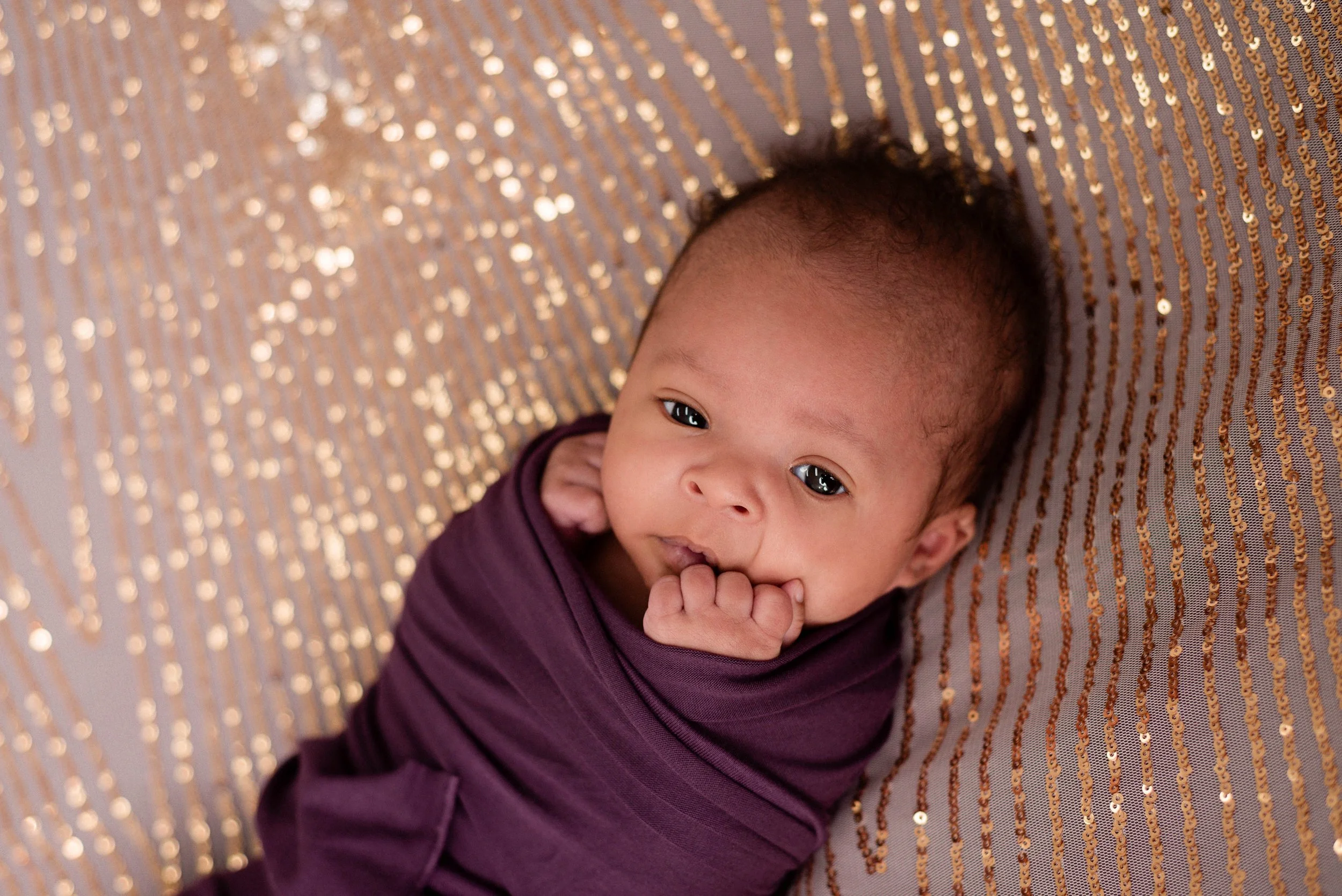 a dark skinned baby with curly hair is swaddled in purple fabric and resting on silver and gold sequined fabric